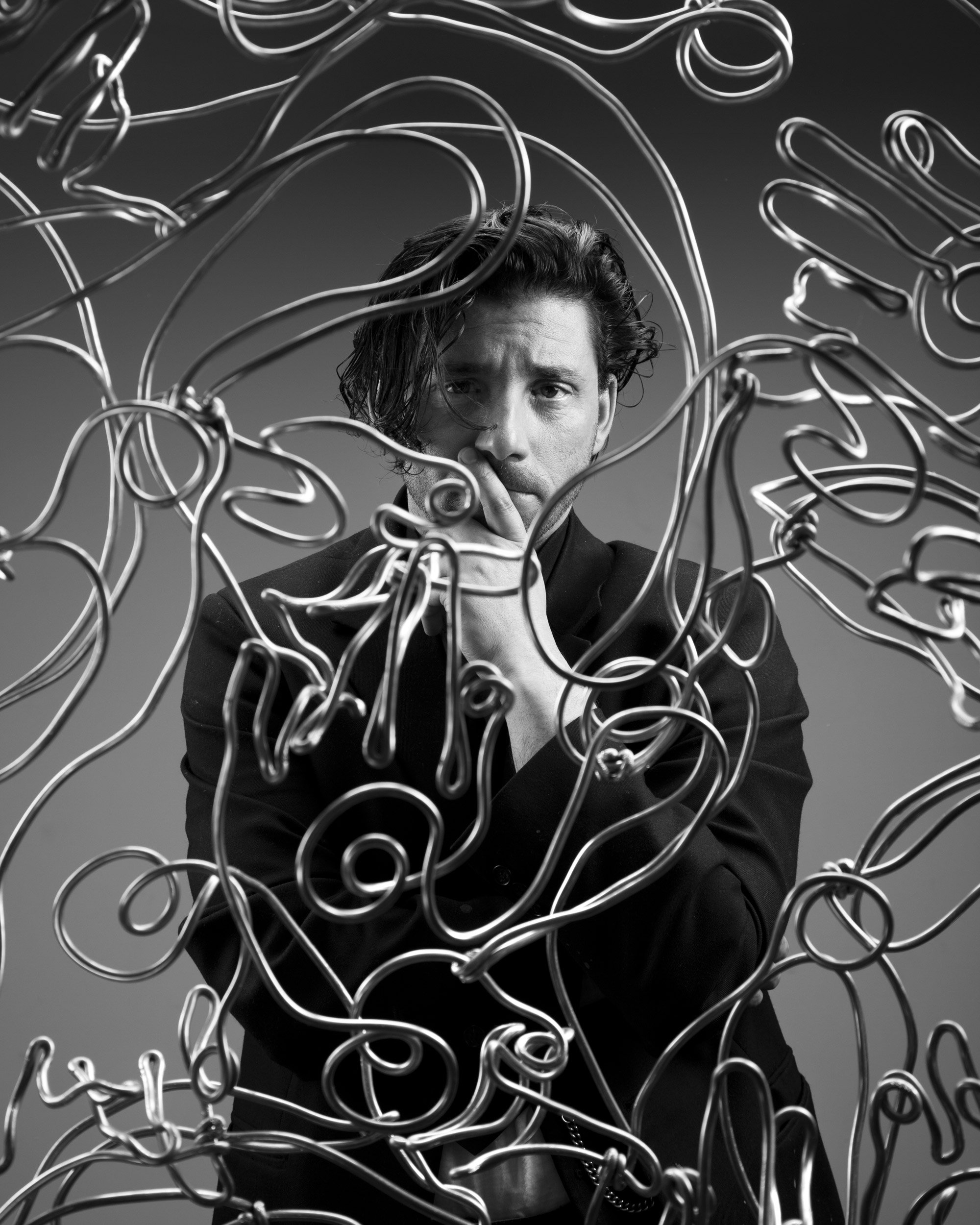 Black and white photograph of a man with wavy hair looking through tangled metal wire sculptures, with cutting face expression, wearing a blazer and a necklace.