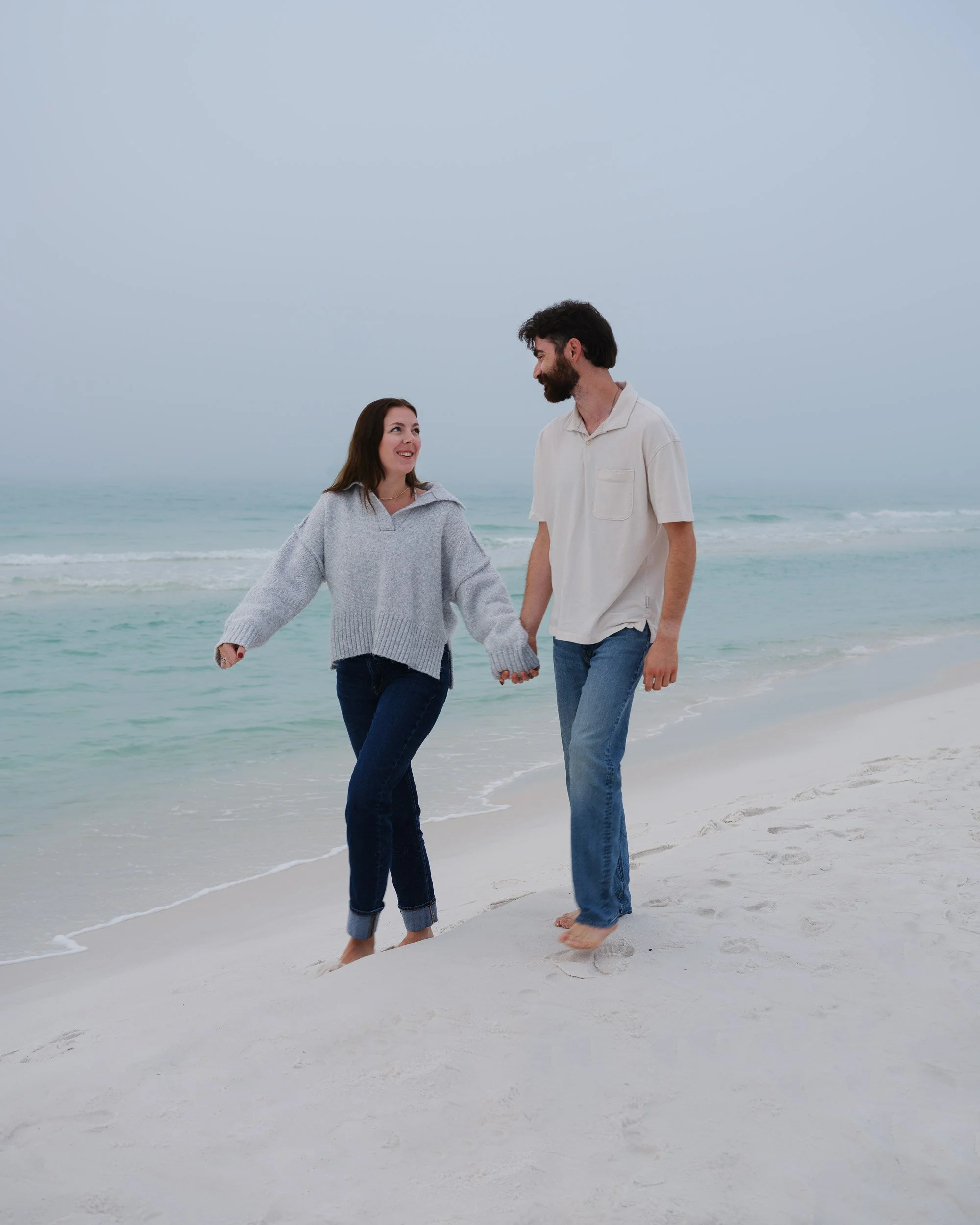 A couple holding hands and walking barefoot on a beach with waves and overcast sky.