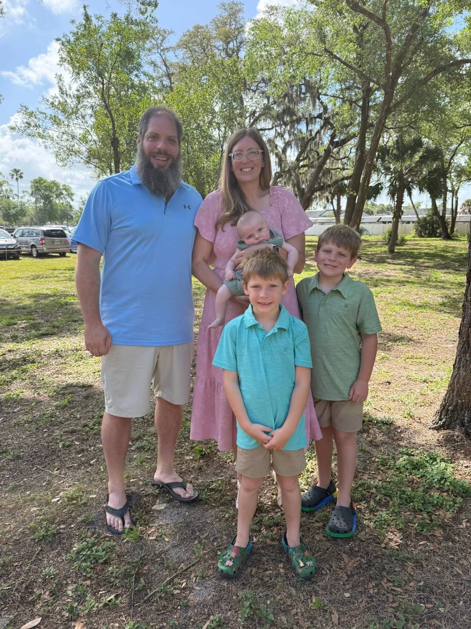 A family photo taken outdoors with five members: a man with a beard in a blue polo shirt, a woman with glasses in a pink dress holding a baby, and three young boys standing in front of them, all under a large tree with cars parked in the background.