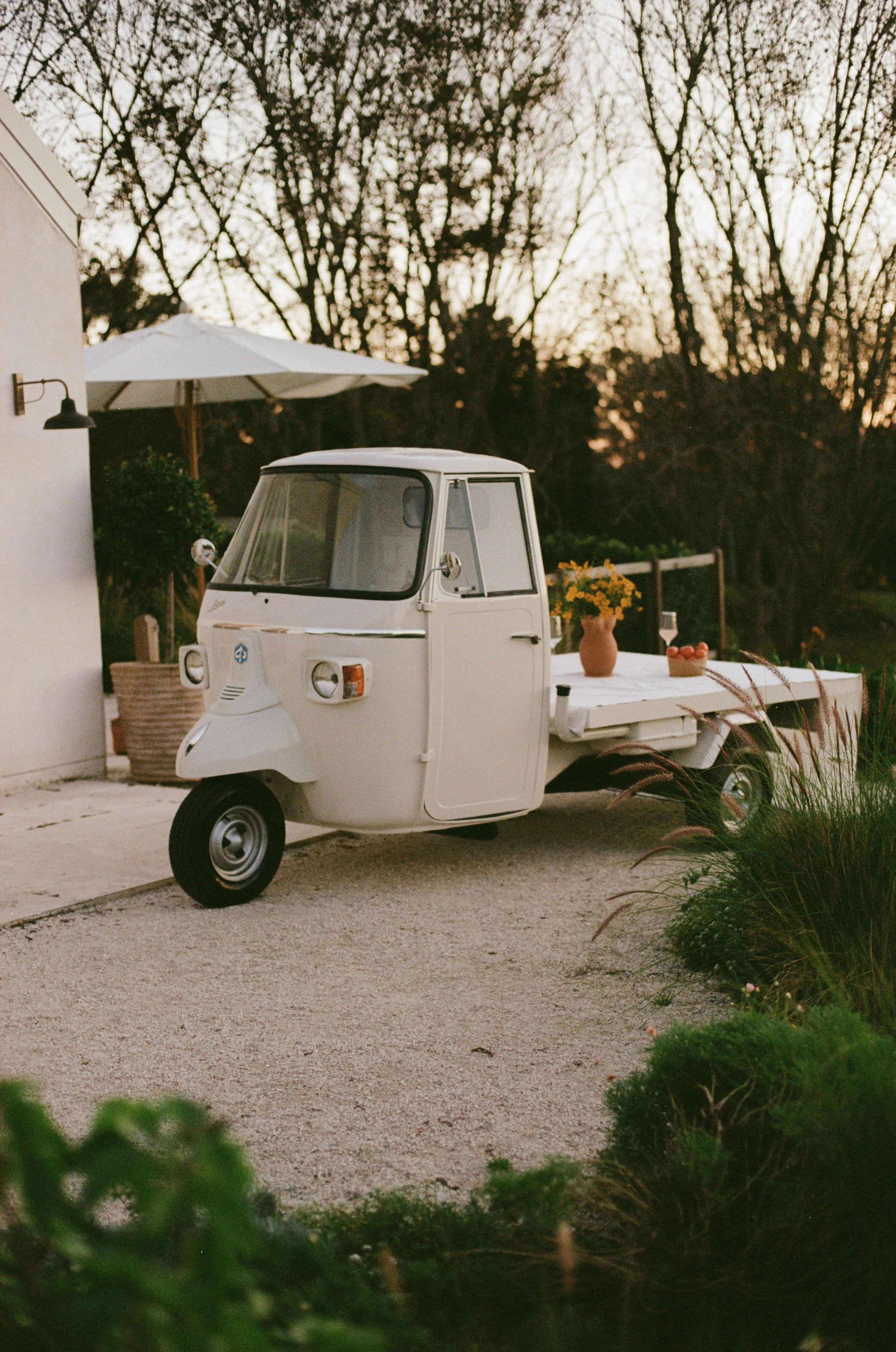 A white motorized Piaggio Ape parked on a gravel driveway near a white building with outdoor furniture and potted plants, with leafless trees in the background at dusk.