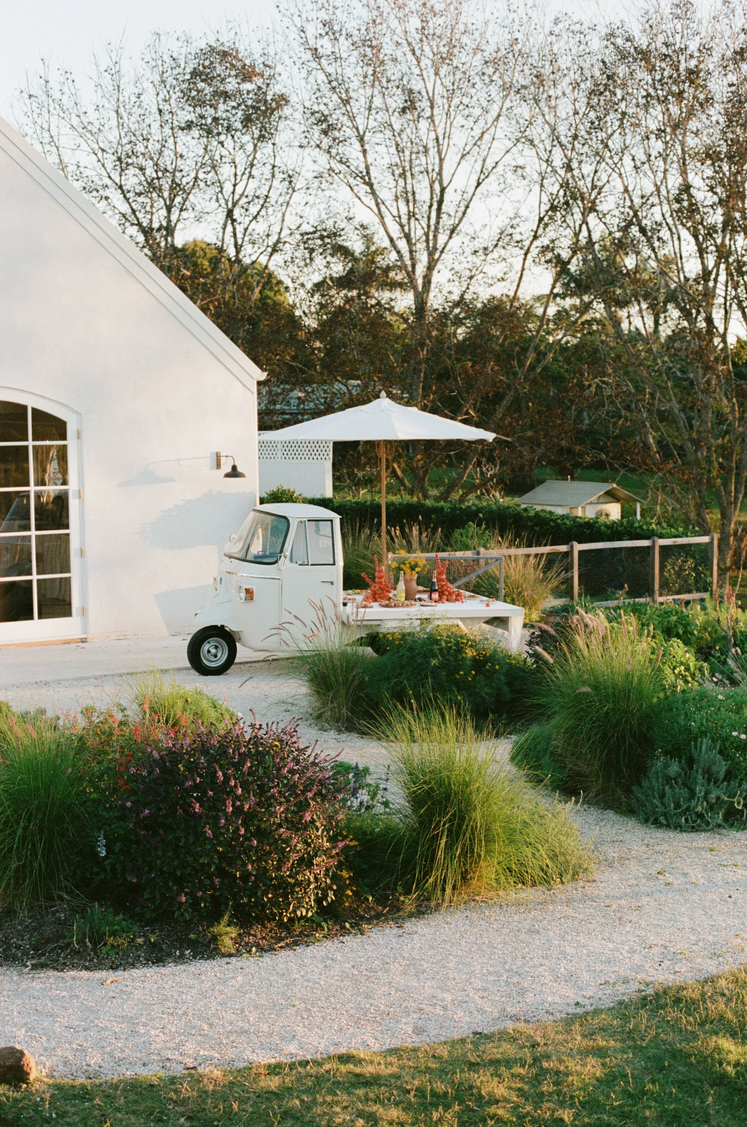 A white house with a large glass door, a small white mini-truck, and an outdoor patio with a white umbrella, surrounded by colorful plants and a garden with a gravel pathway. There are trees in the background, and the image is captured at sunset.