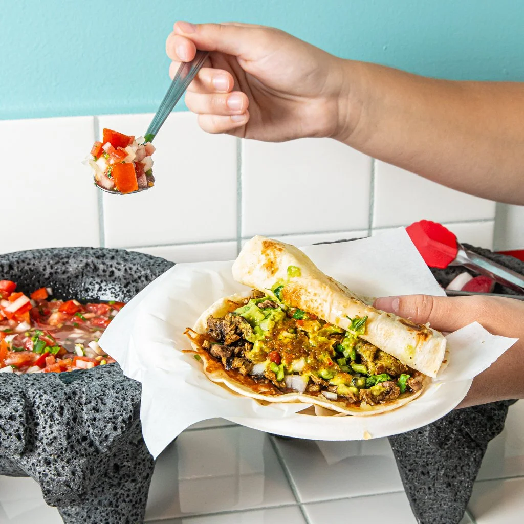 Person holding a taco filled with beef, lettuce, and salsa, holding a spoonful of pico de gallo above the taco in a kitchen setting. Food Photography Toronto.