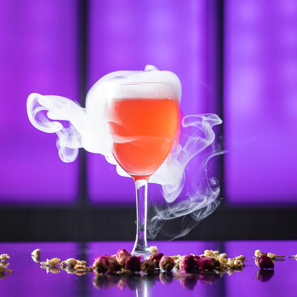 A glass of pink cocktail with white smoke or vapor swirling around it, on a reflective surface with flower petals scattered around, against a purple background. Cocktail Photography Toronto.