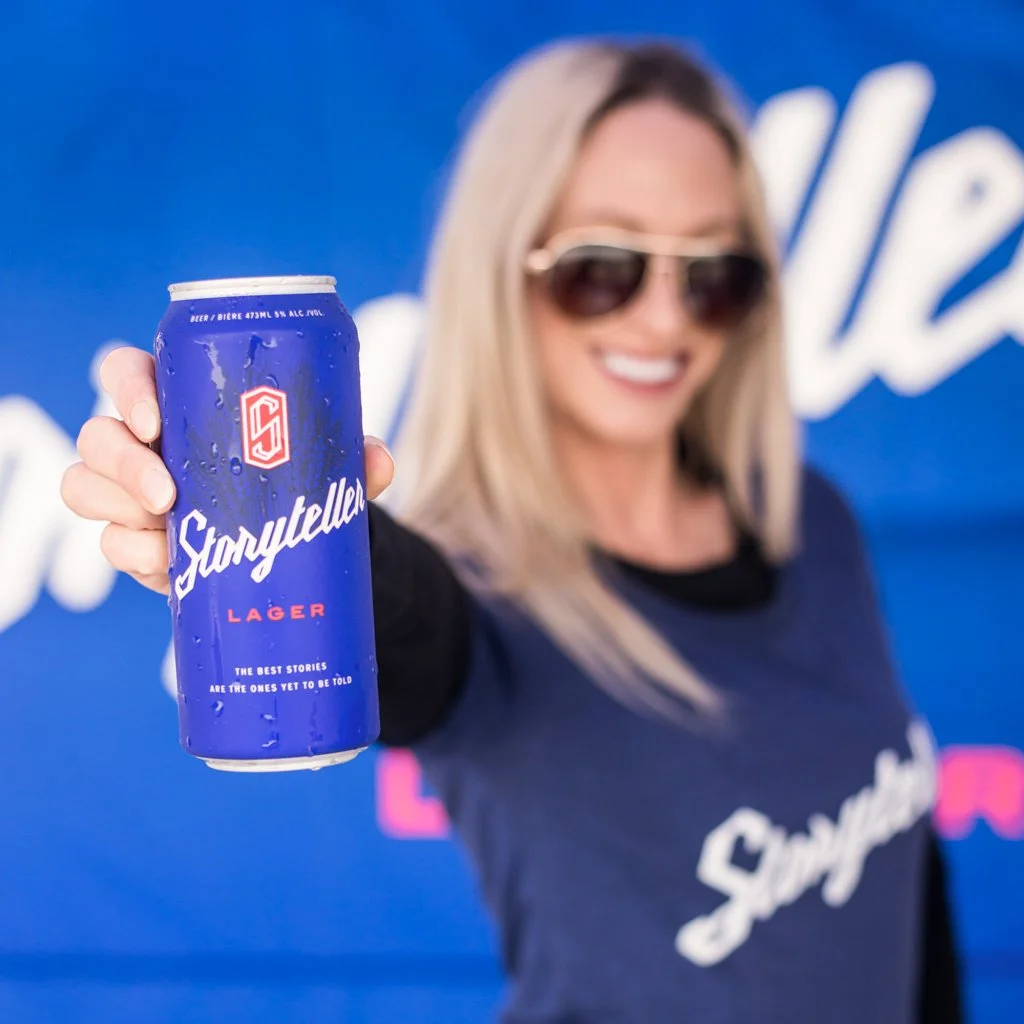 A woman with blonde hair wearing sunglasses and a navy blue t-shirt, holding a blue can of Starry Teller Lager beer towards the camera with a blue backdrop. Food Event Photography Toronto.