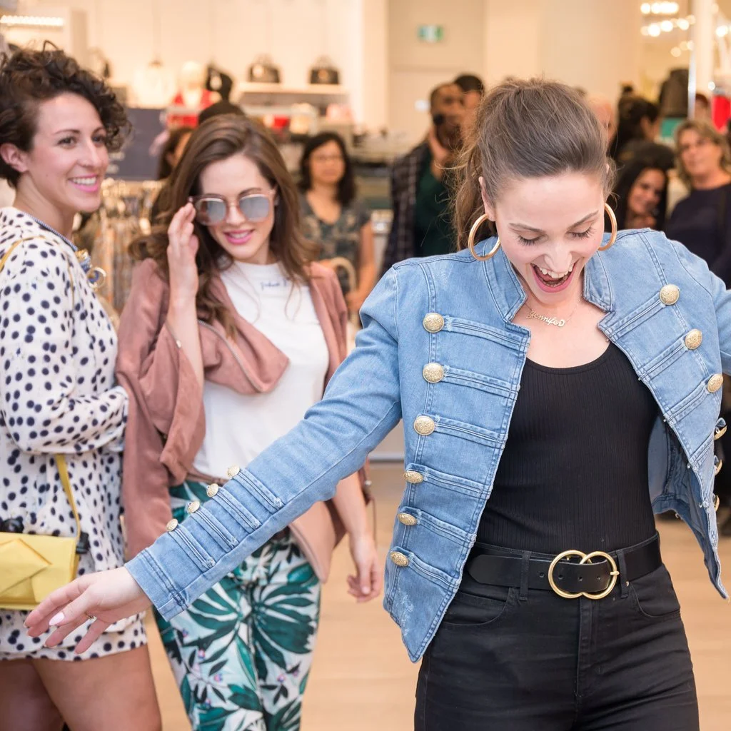 A woman in a denim jacket and black pants laughs while dancing in a crowded store, with two women smiling nearby. Brand Photographer Toronto. Event Photographer Toronto.