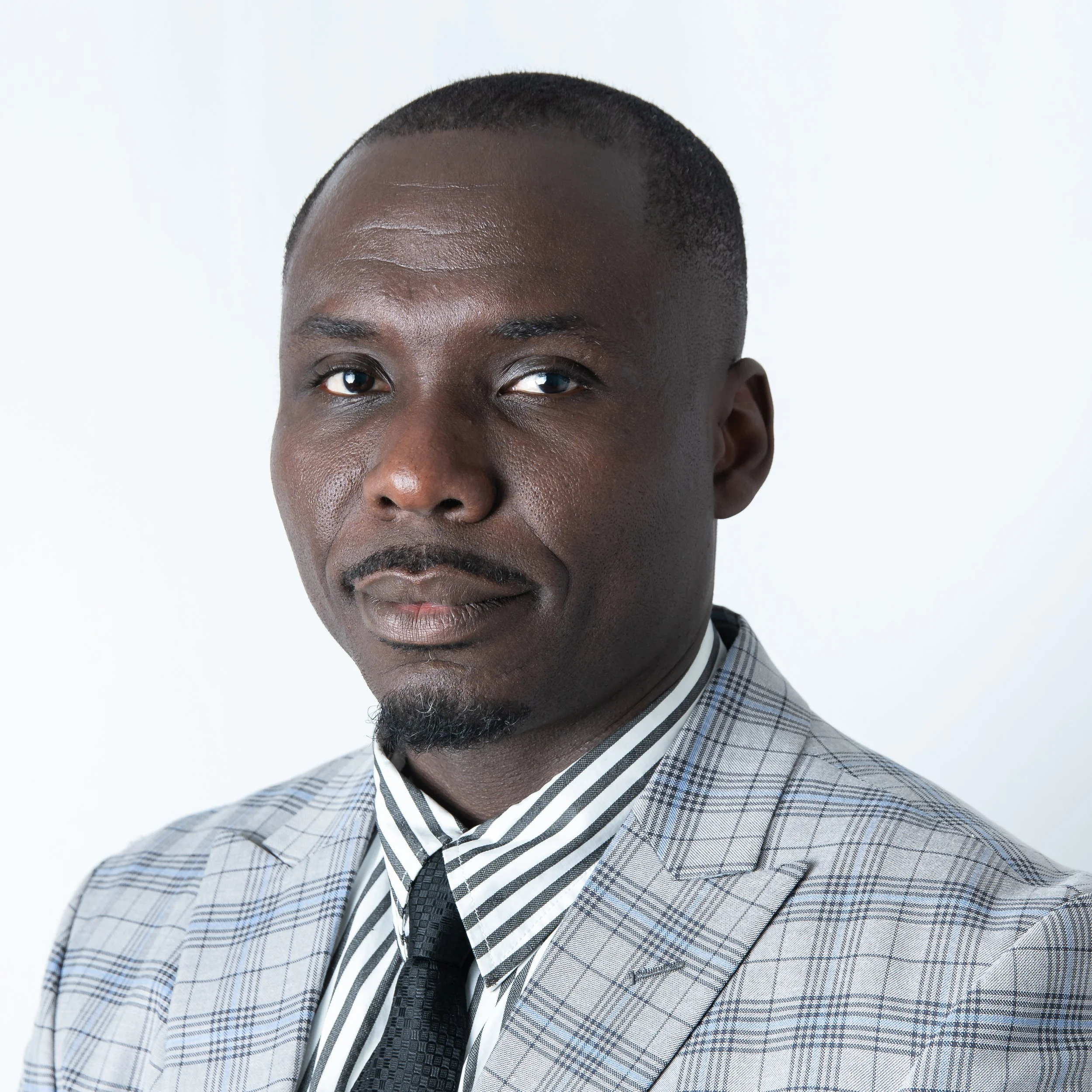 Close-up of a man's face with dark skin, short hair, and a part of his checkered suit visible, against a plain white background. Headshot Photographer Toronto. Lawyer Headshots Toronto.