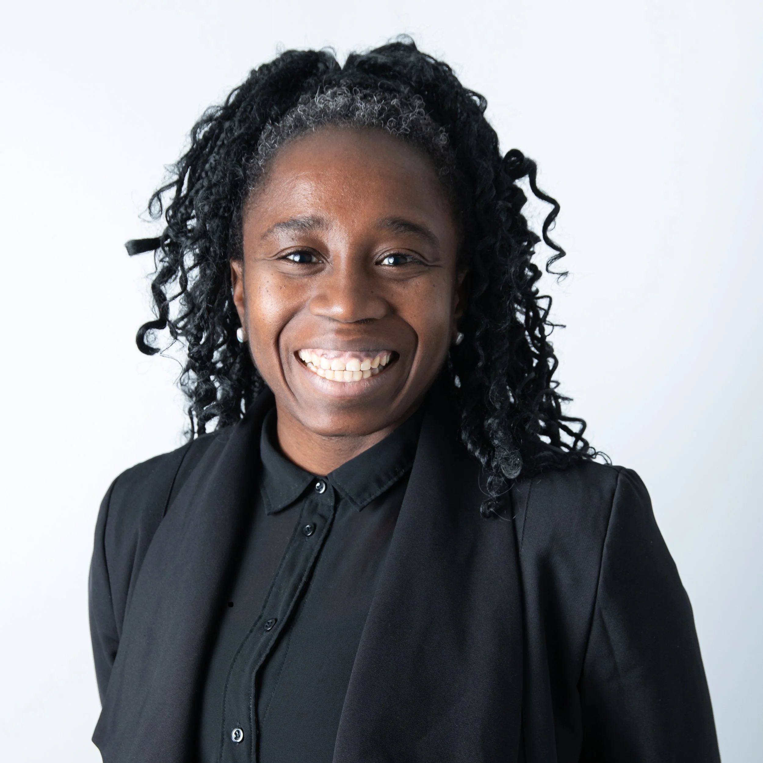 Close-up of a woman with dark skin, curly black hair styled in twists, wearing a black button-up shirt, smiling against a plain white background. Headshot Photographer Toronto. Lawyer Headshots Toronto.