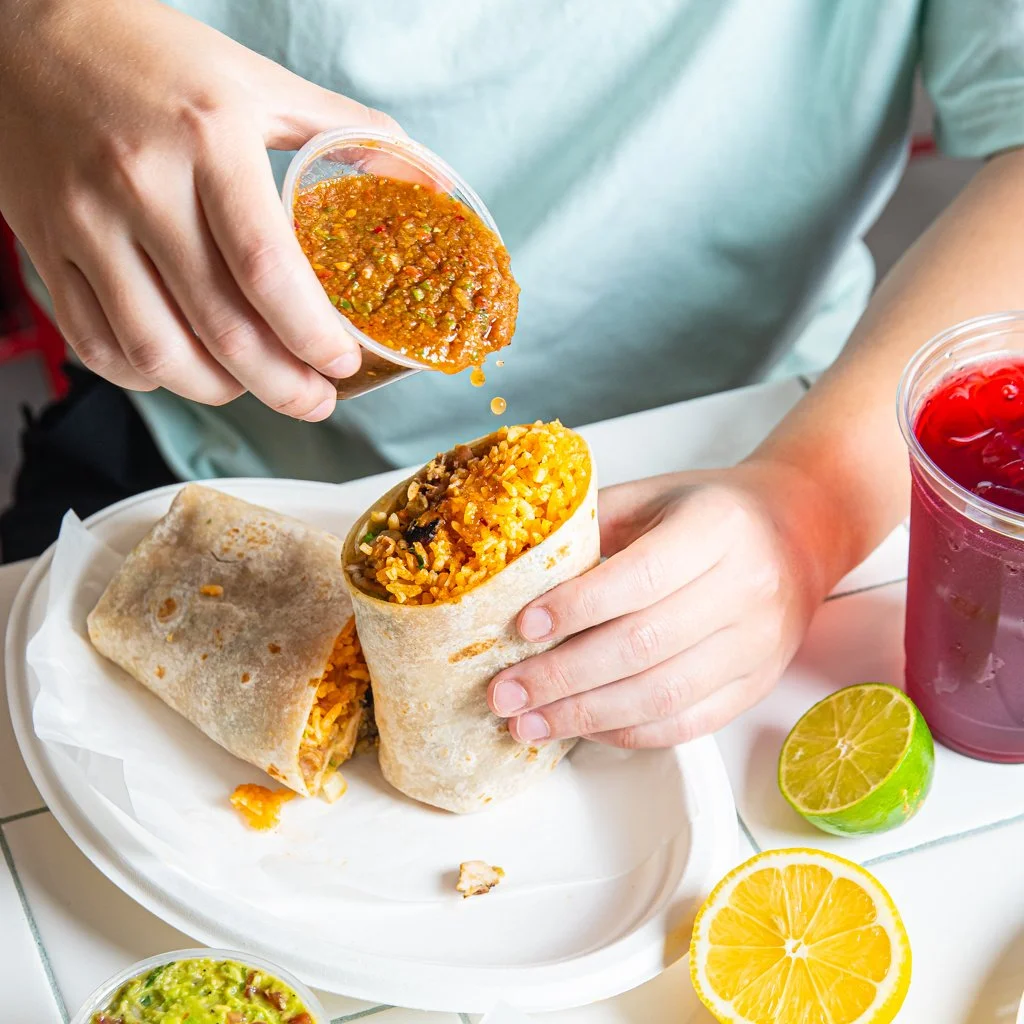 Person holding a burrito with rice and vegetables, pouring salsa over it, with a lime half and a lemon slice on the table, alongside a drink with red liquid and ice. Food Photography Toronto.