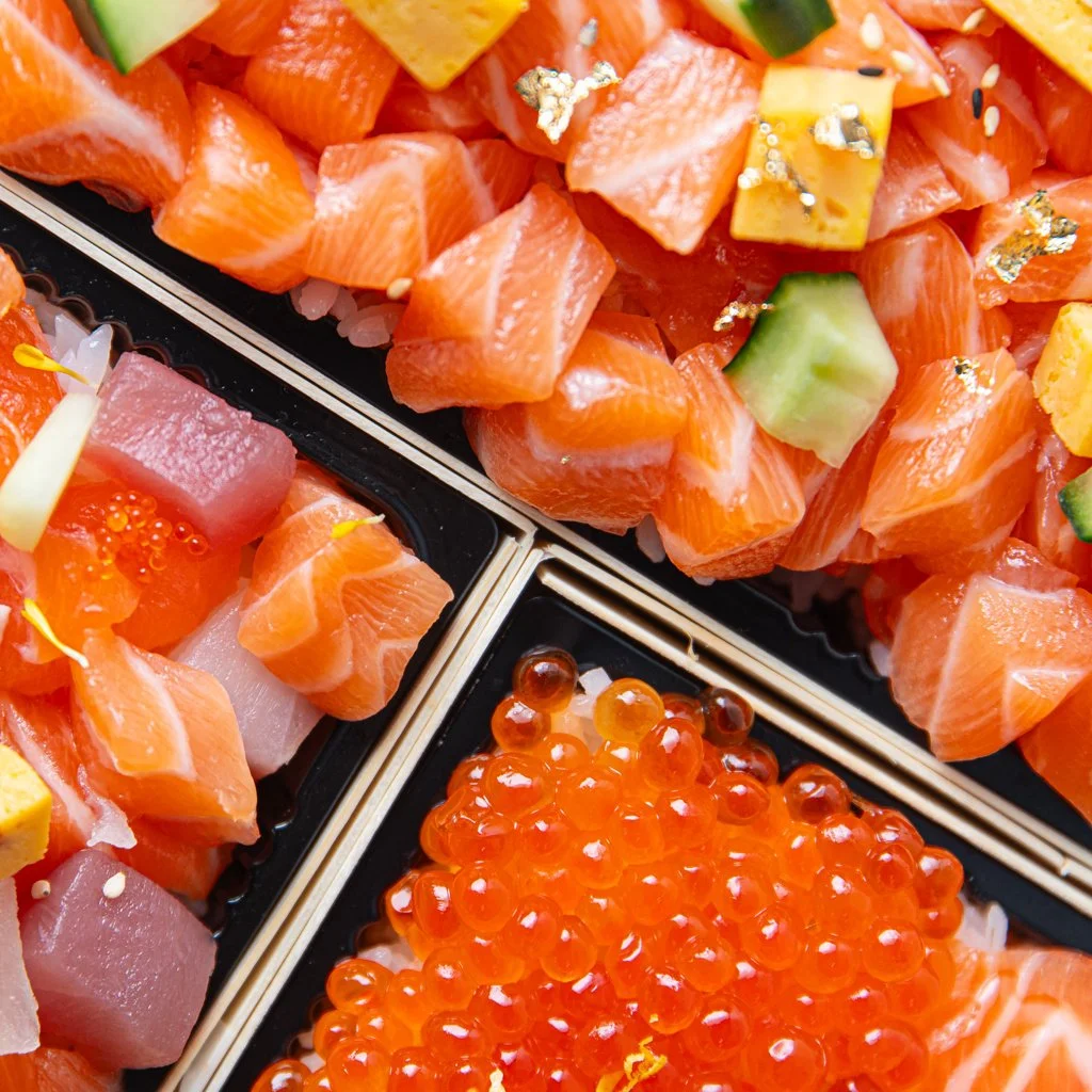 Close-up of assorted sashimi, including salmon, tuna, and yellowtail, topped with cucumber, gold leaf, and placed on rice, with bright orange salmon roe. Sushi Photography Toronto
