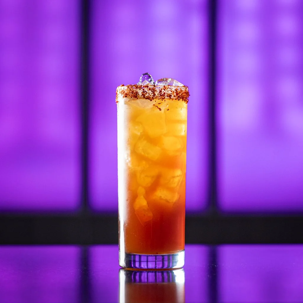 A tall glass of iced cocktail garnished with a red chili powder rim and a purple flower, set against a purple illuminated background. Cocktail Photography Toronto.