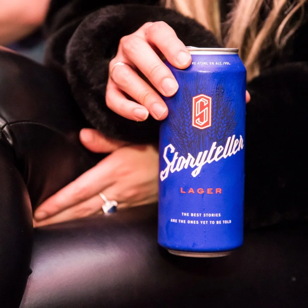 A person holding a blue can of Storyteller Lager beer with one hand, with a ring on their finger, resting on a black surface. Food Event Photography Toronto.