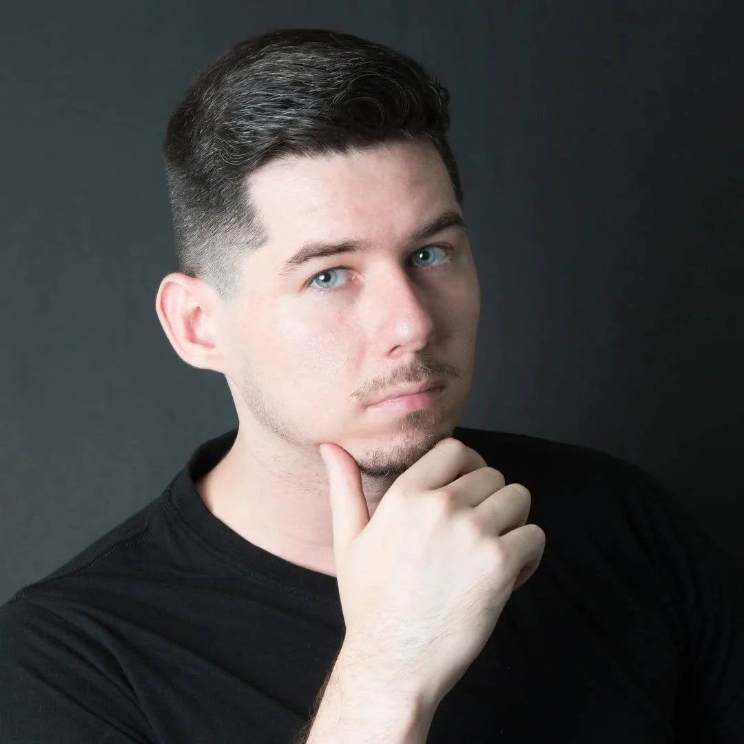 Young man with short brown hair and blue eyes, wearing a black shirt, holding his chin thoughtfully against a dark background.