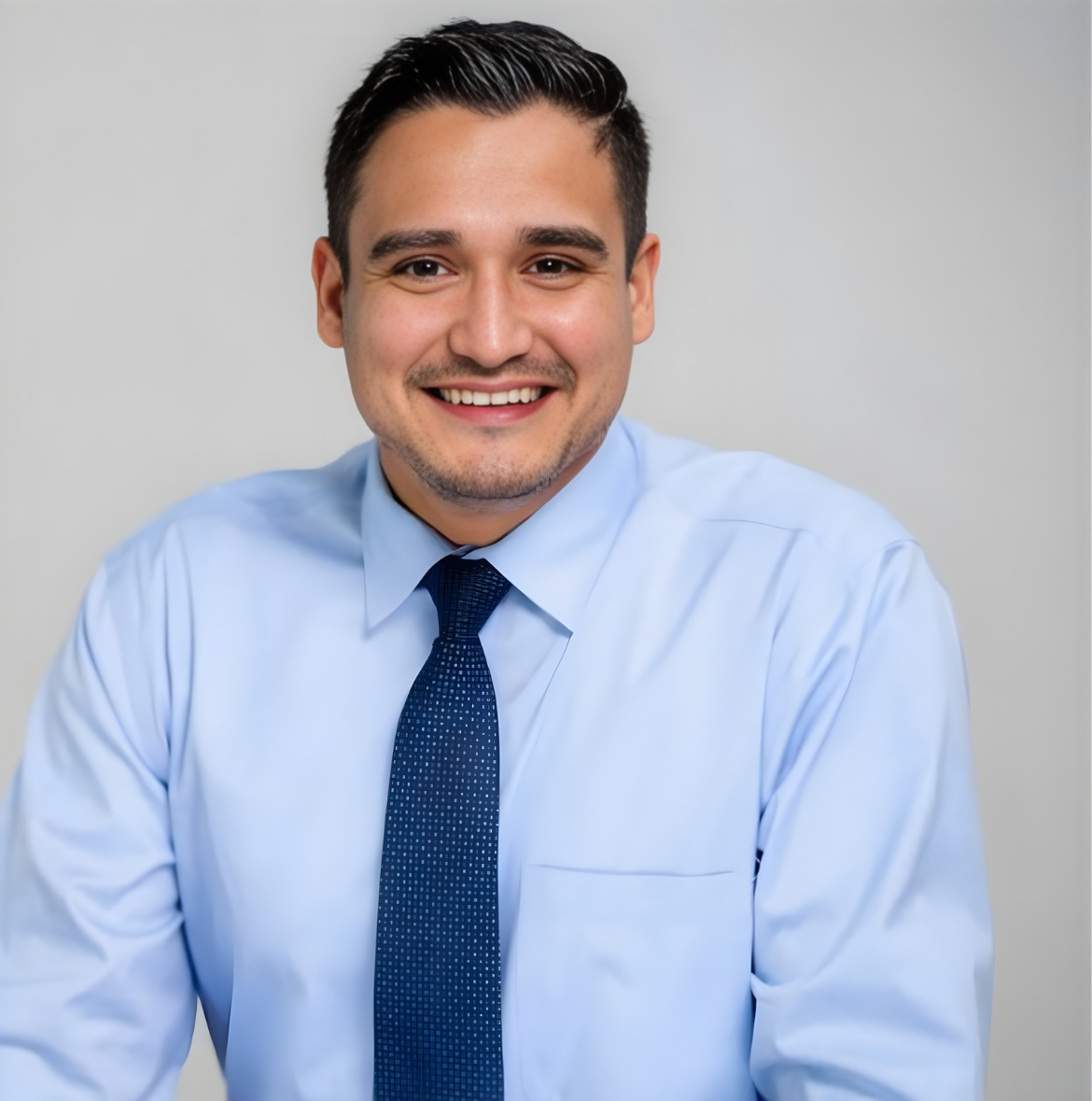 A smiling young man in a light blue shirt and dark blue tie, sitting against a plain light gray background.