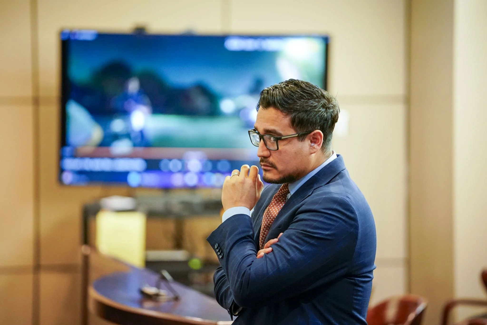 A man with dark curly hair and glasses, dressed in a blue suit and orange patterned tie, sits with his eyes closed and hand near his face in a conference room, with a large TV screen in the background.