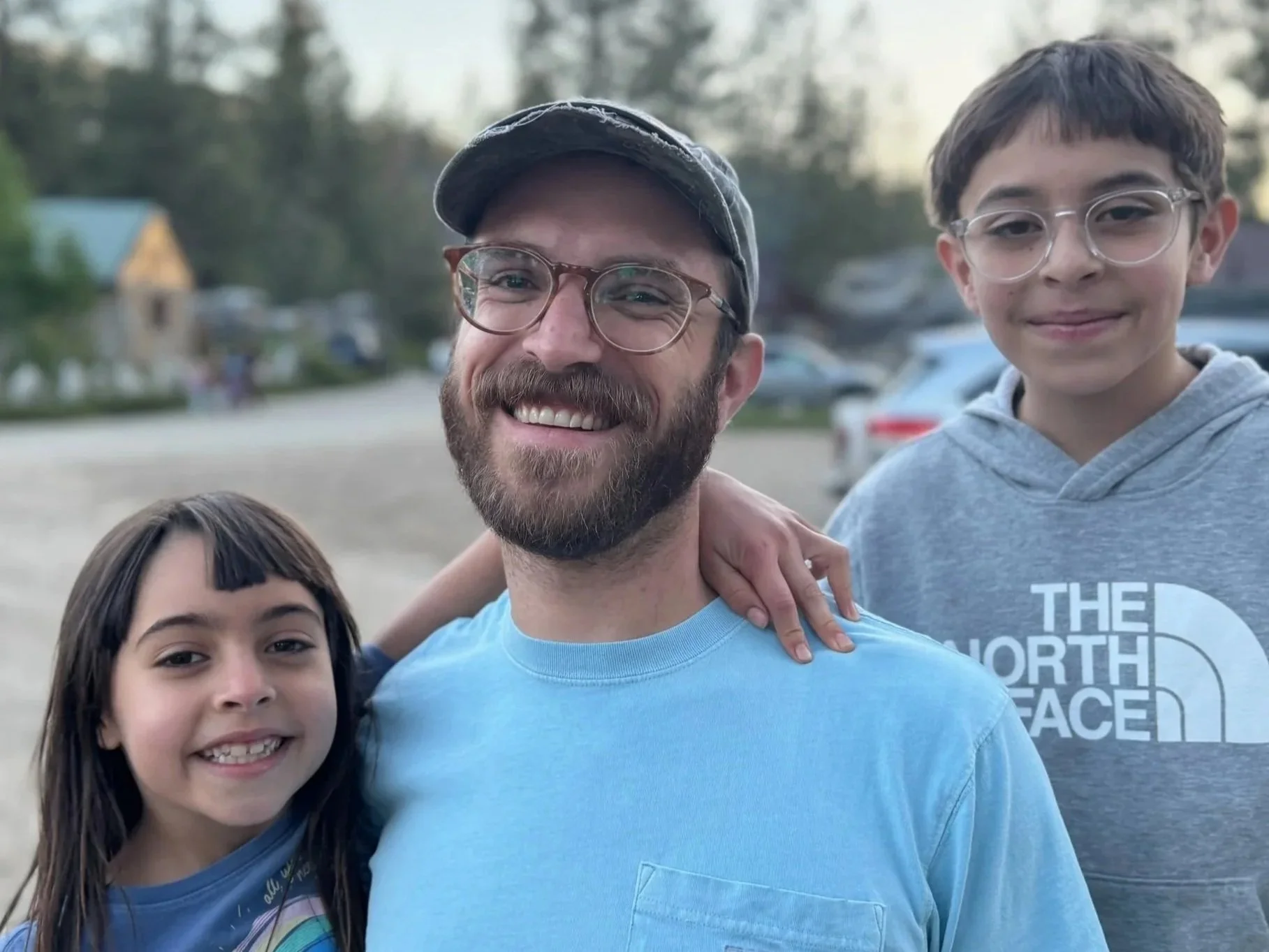 A photo of a smiling family, including a man with glasses and a beard, a young boy with glasses, and part of a girl's face and shoulder, standing outdoors in a park or open area with trees and parked cars in the background.