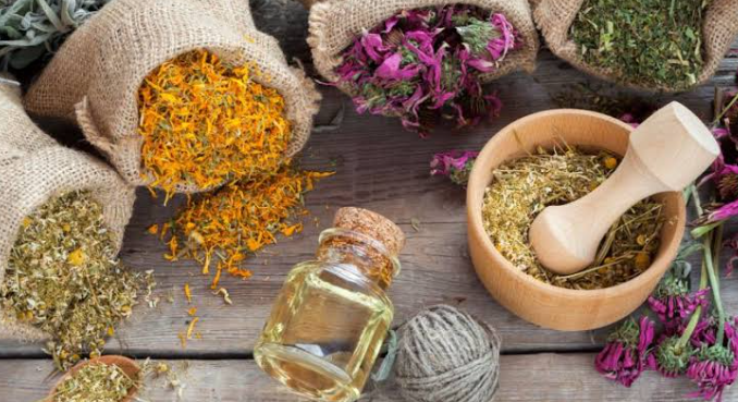 Hierbas secas y flores, un mortero con un mazo, un frasco de aceite, y una bola de hilo en una mesa de madera.