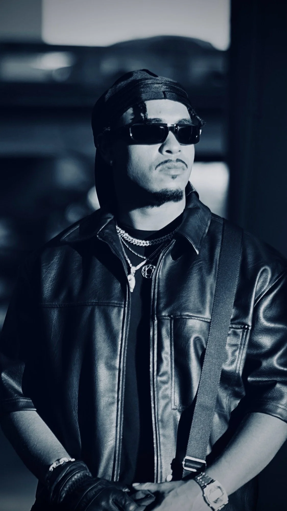 A person wearing sunglasses, a bandana, multiple necklaces, a leather jacket, and a watch, looking to the side in a black-and-white photo.