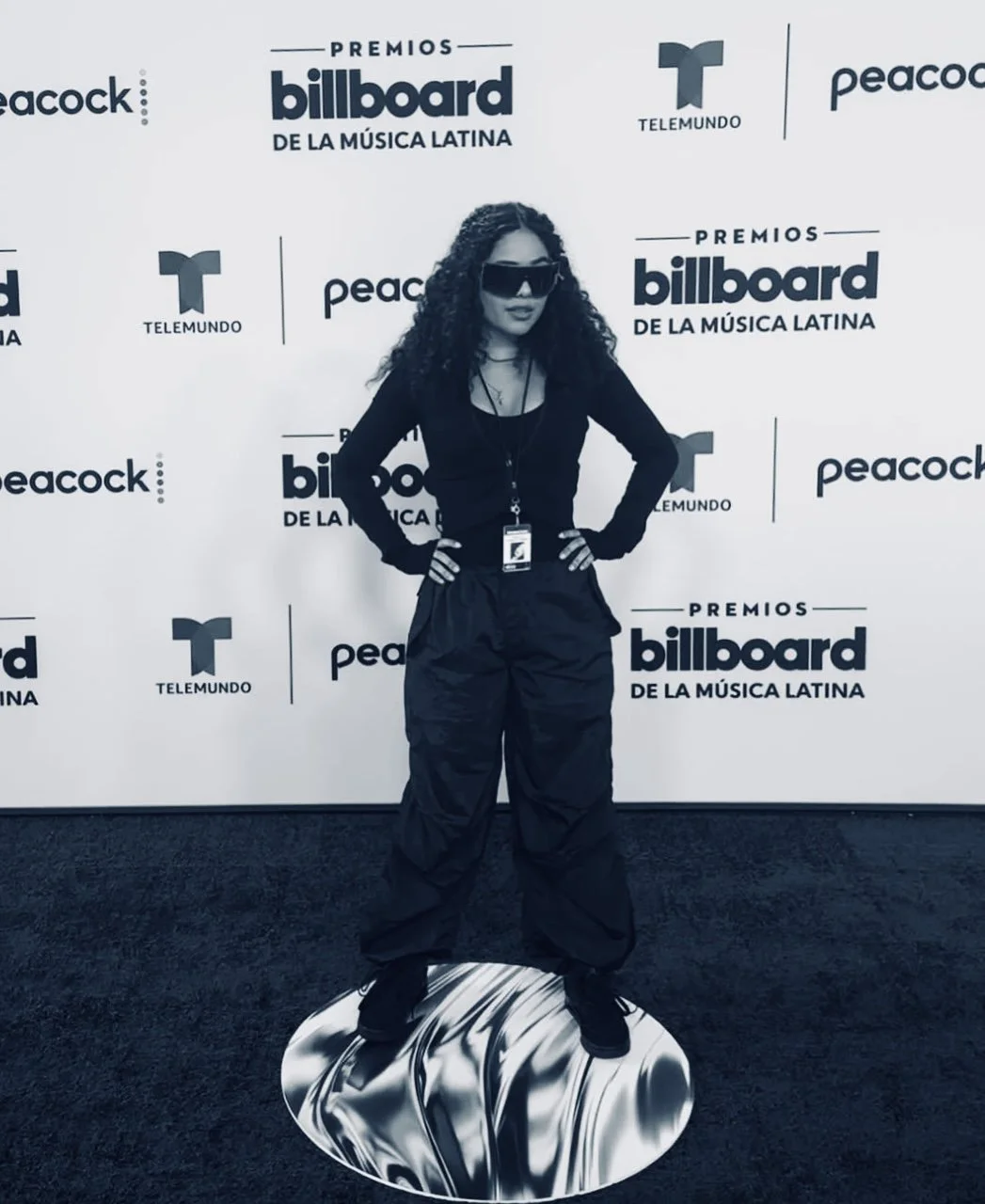 A woman with curly hair wearing sunglasses, a black long-sleeve top, baggy black pants, and sneakers, standing with hands on hips in front of a backdrop with logos of Billboard Latin Music Awards, Telemundo, and Peacock. She is standing on a reflective, metallic-looking platform.