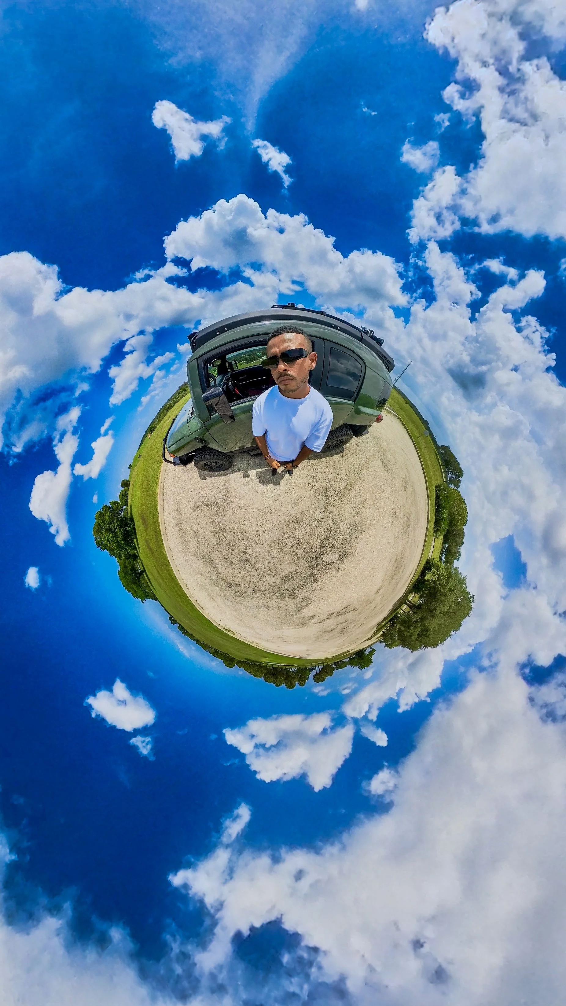 A man wearing sunglasses and a white t-shirt standing next to a green vehicle on a round, dirt-covered area with green grass and trees around. The sky is bright blue with scattered white clouds, and the photo appears to be taken with a 360-degree camera creating a tiny planet effect.