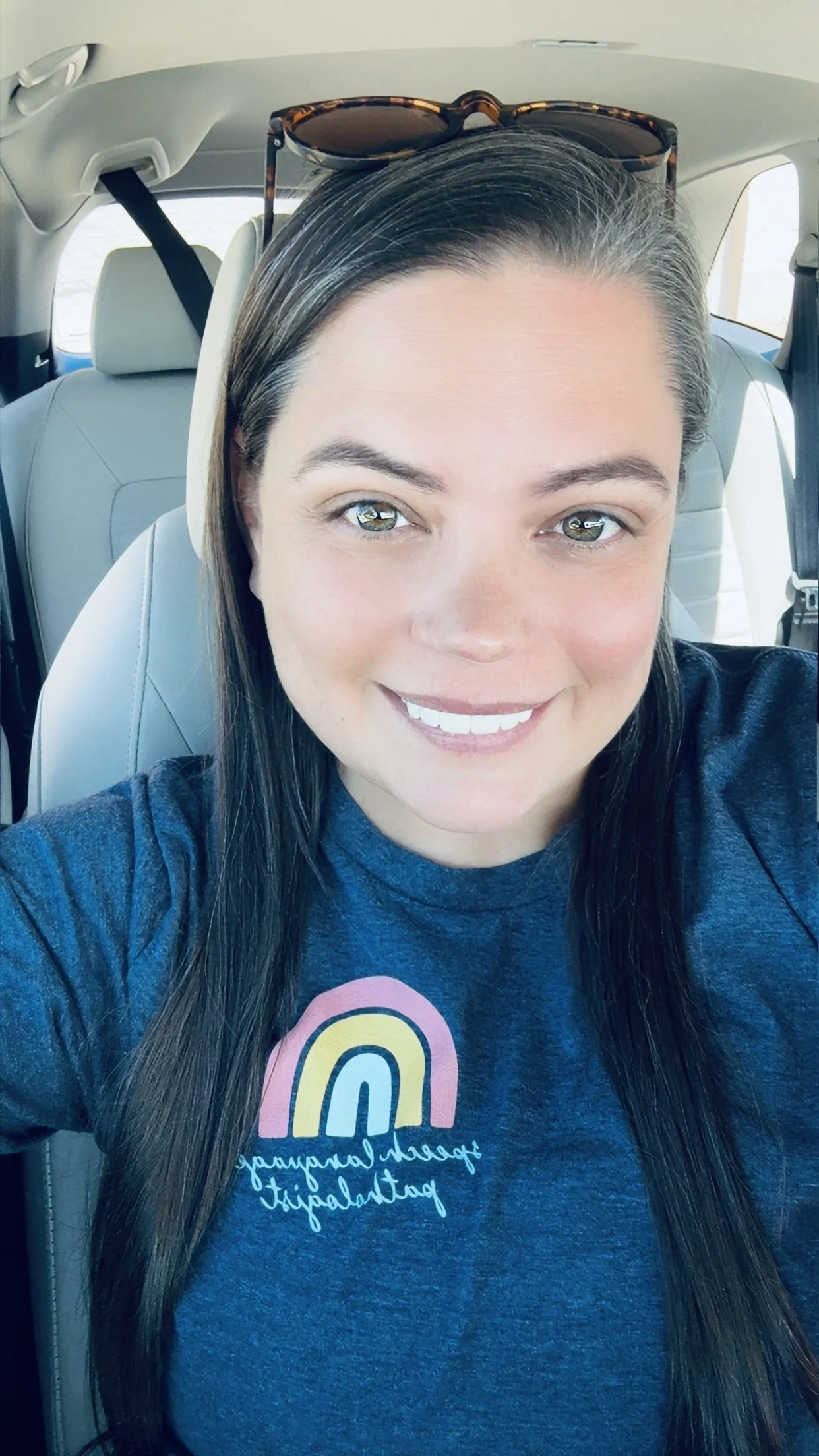 A woman with long dark hair and glasses resting on her head, smiling for a selfie inside a vehicle, wearing a dark blue T-shirt with colorful rainbow graphics.