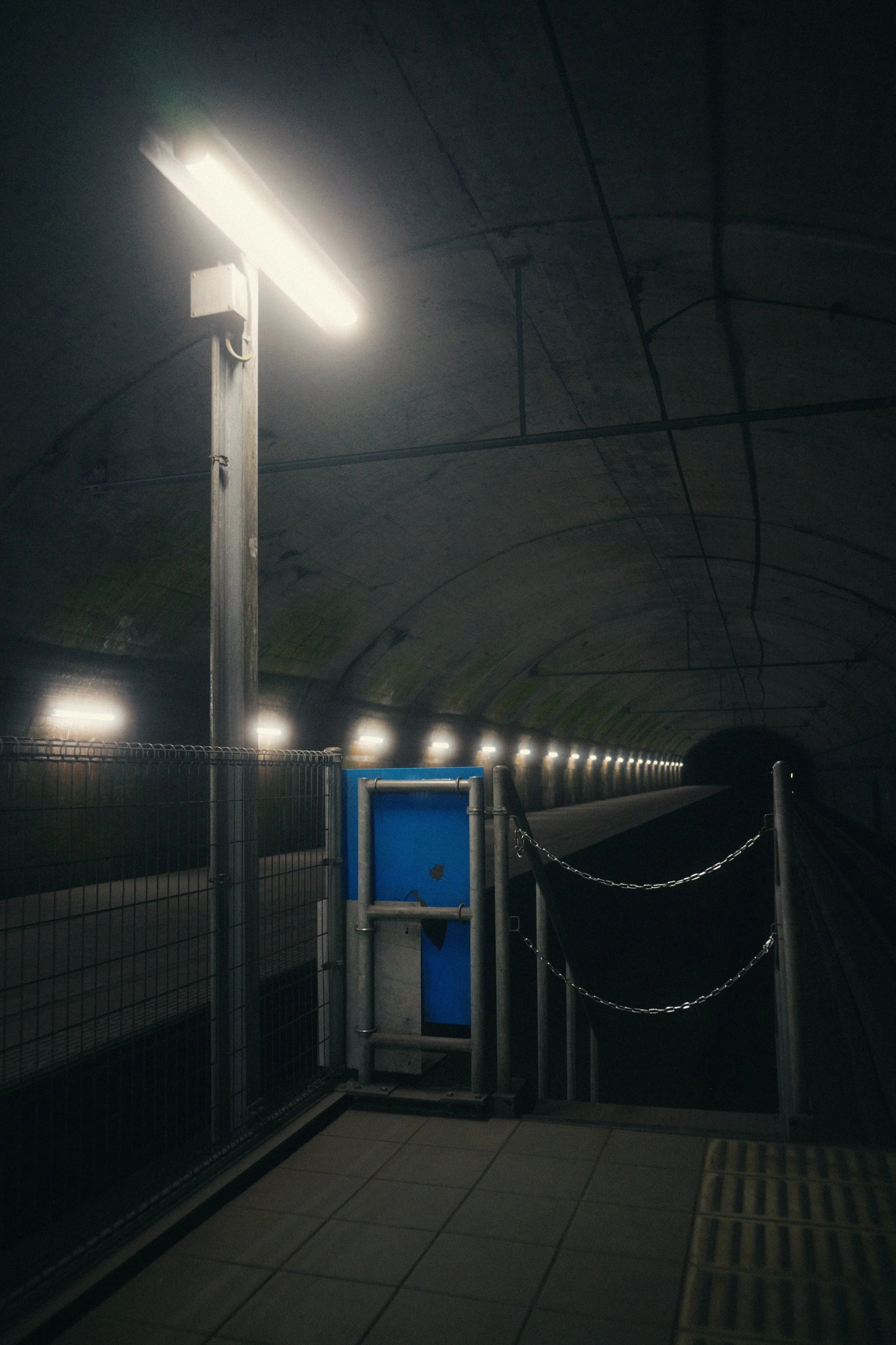 View of a dark tunnel with concrete walls and ceiling, illuminated by fluorescent lights along the ceiling. In the foreground, there is a fenced-off area with a gate and a blue informational sign, and the tunnel extends into darkness.