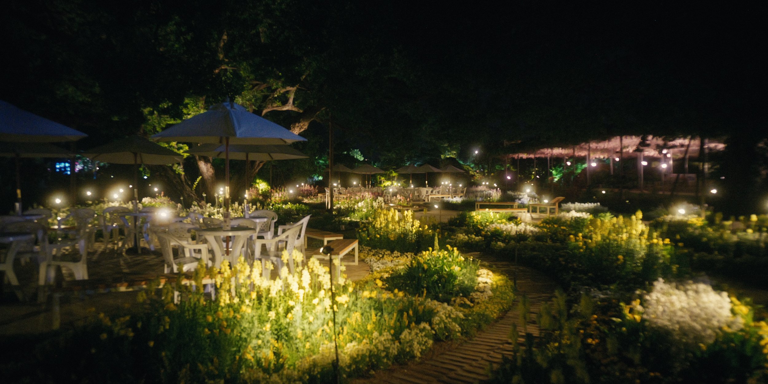 Nighttime outdoor garden with tables, chairs, and umbrellas illuminated by small lights. Flowers line pathways and trees are visible in the background.