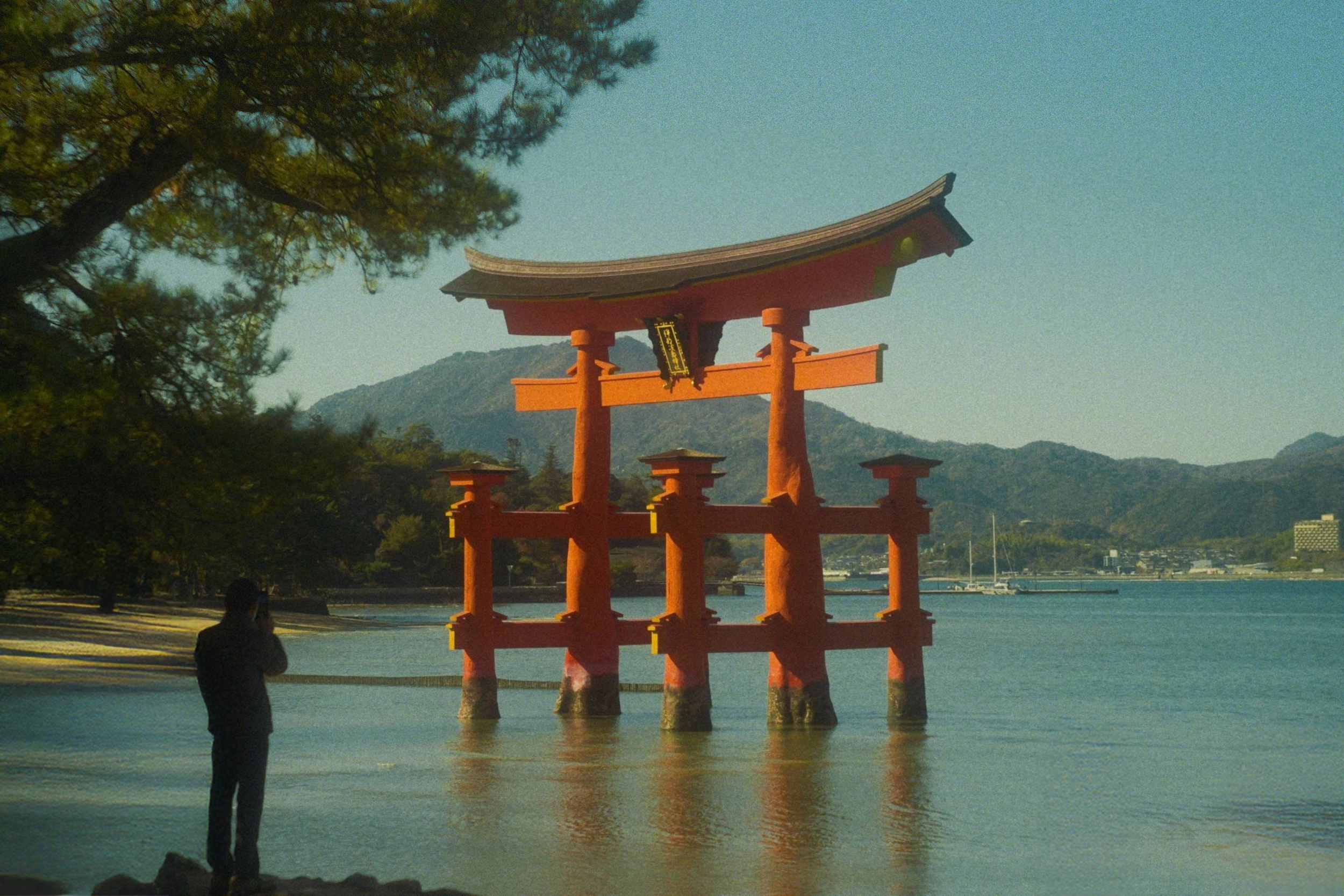 A traditional Japanese torii gate painted in red, partially submerged in water, with trees on the left and a mountain in the background under a clear sky.