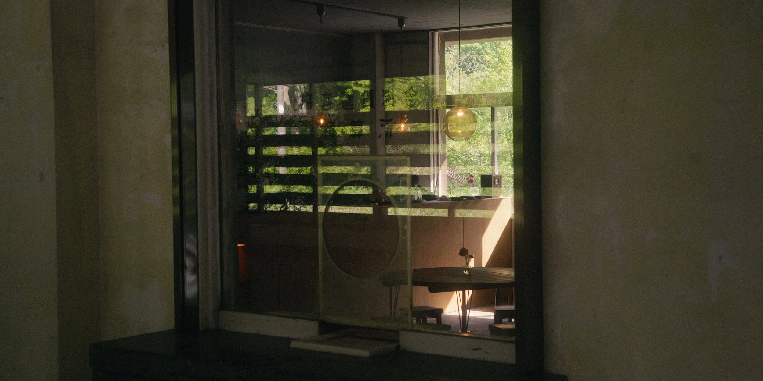 Interior view of a room with large glass windows showing green trees outside, sunlight streaming in, with a desk, chair, and decorative items inside.