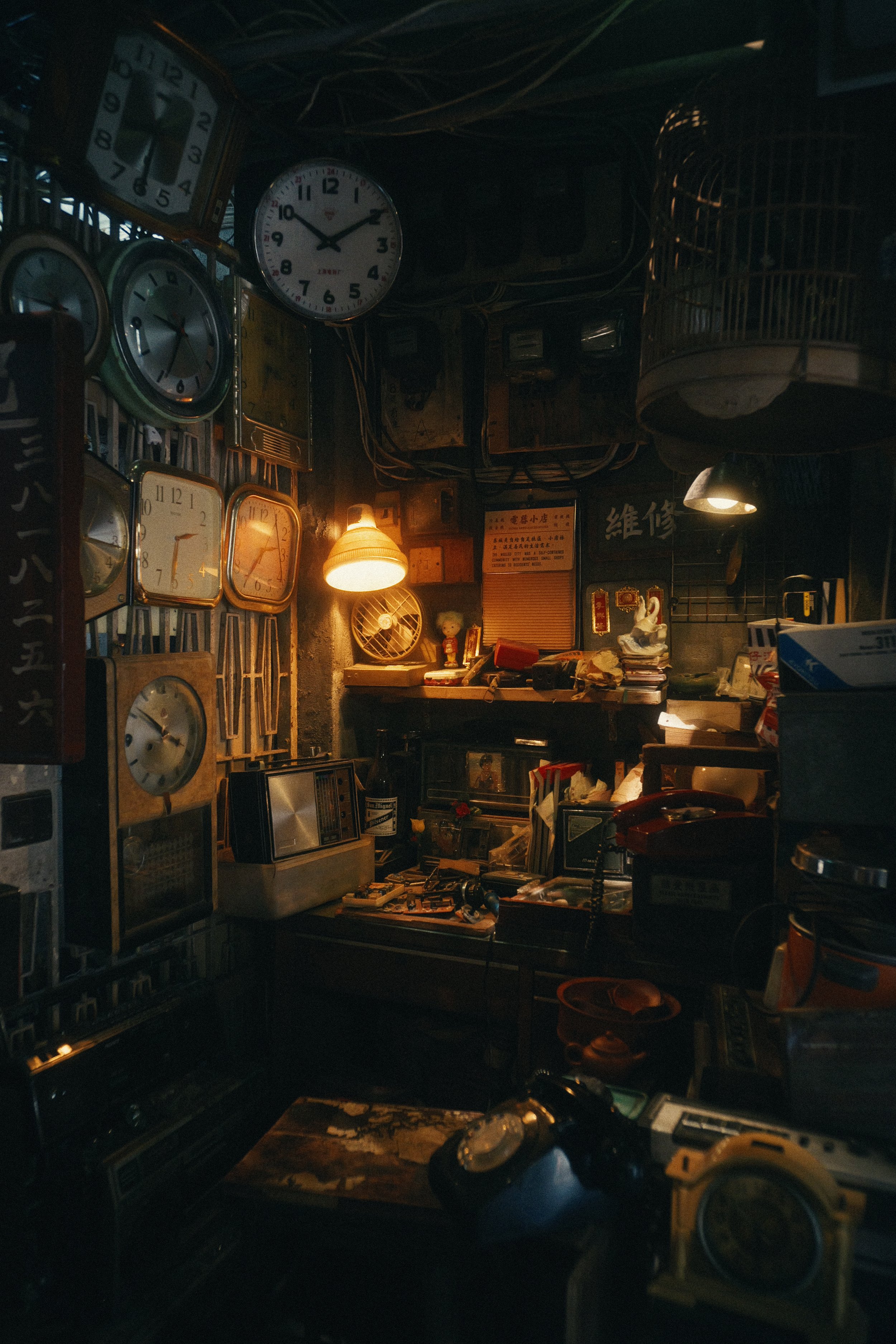 A cluttered room with multiple clocks on the wall, a desk with various objects, a small fan, and warm lighting.