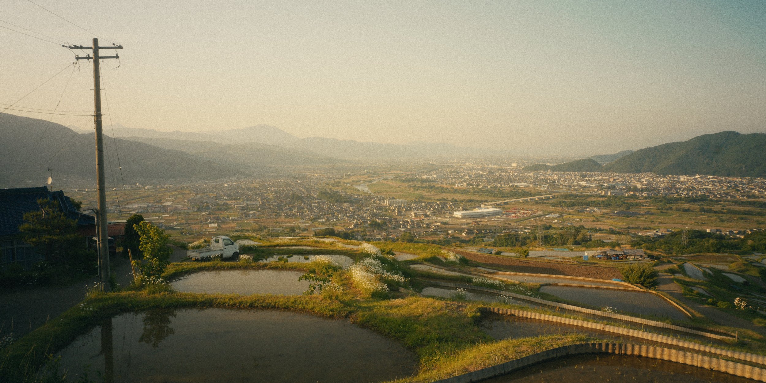 A panoramic view of a city in a valley surrounded by mountains, with rice paddies in the foreground and a utility pole on the left.