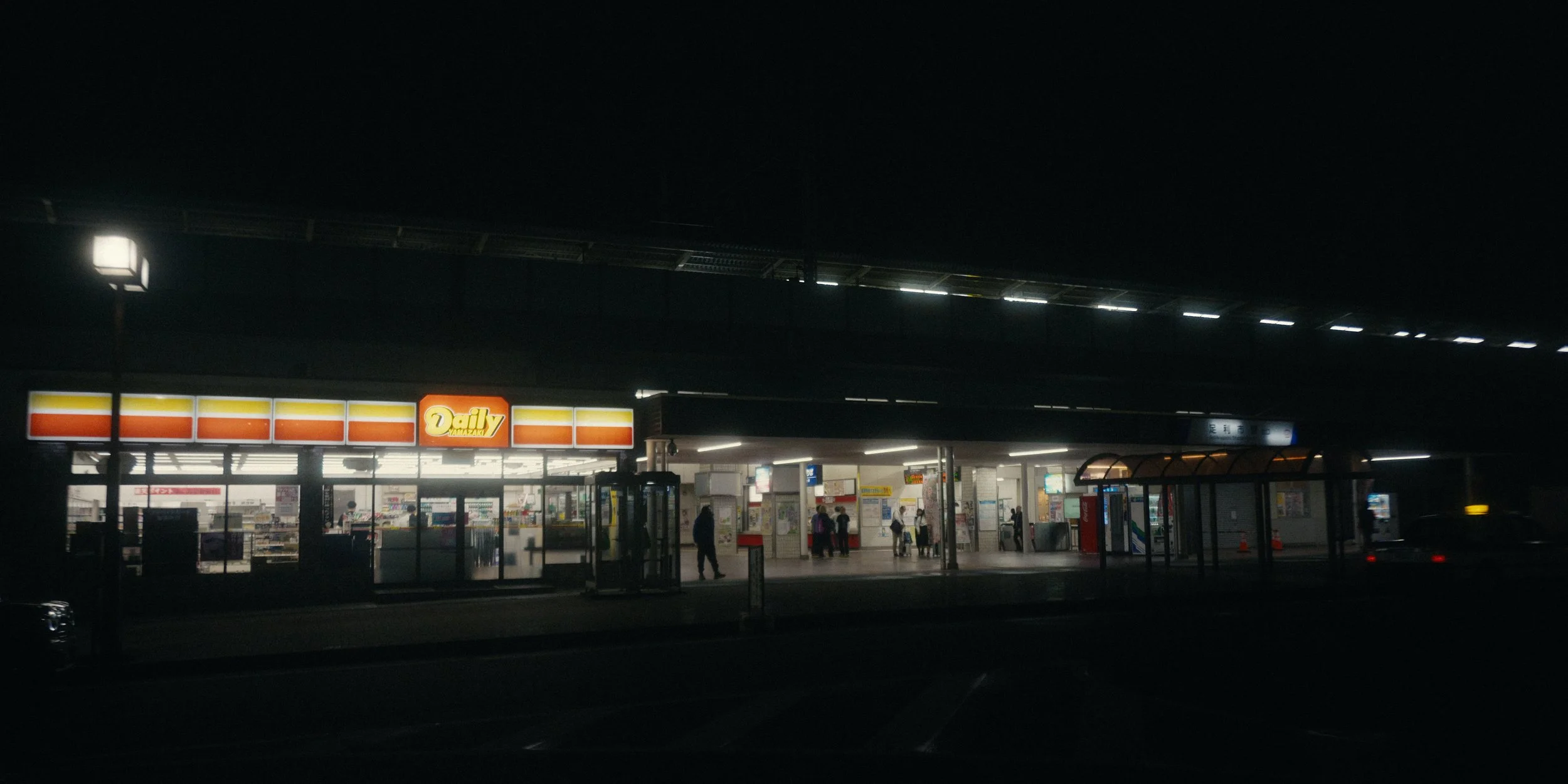 Night view of a convenience store called 'Daily', with people entering and exiting, illuminated signage, and a bus stop in front.
