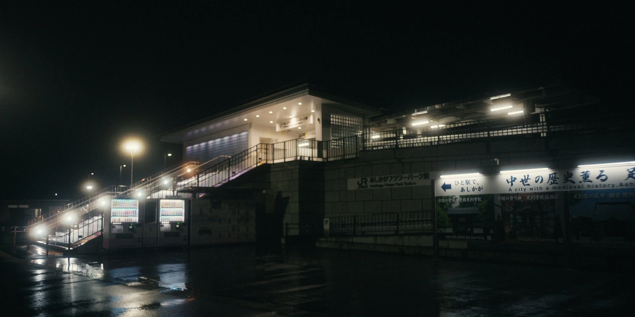Nighttime photo of a train station entrance, illuminated signs, and vending machines outside a modern building.