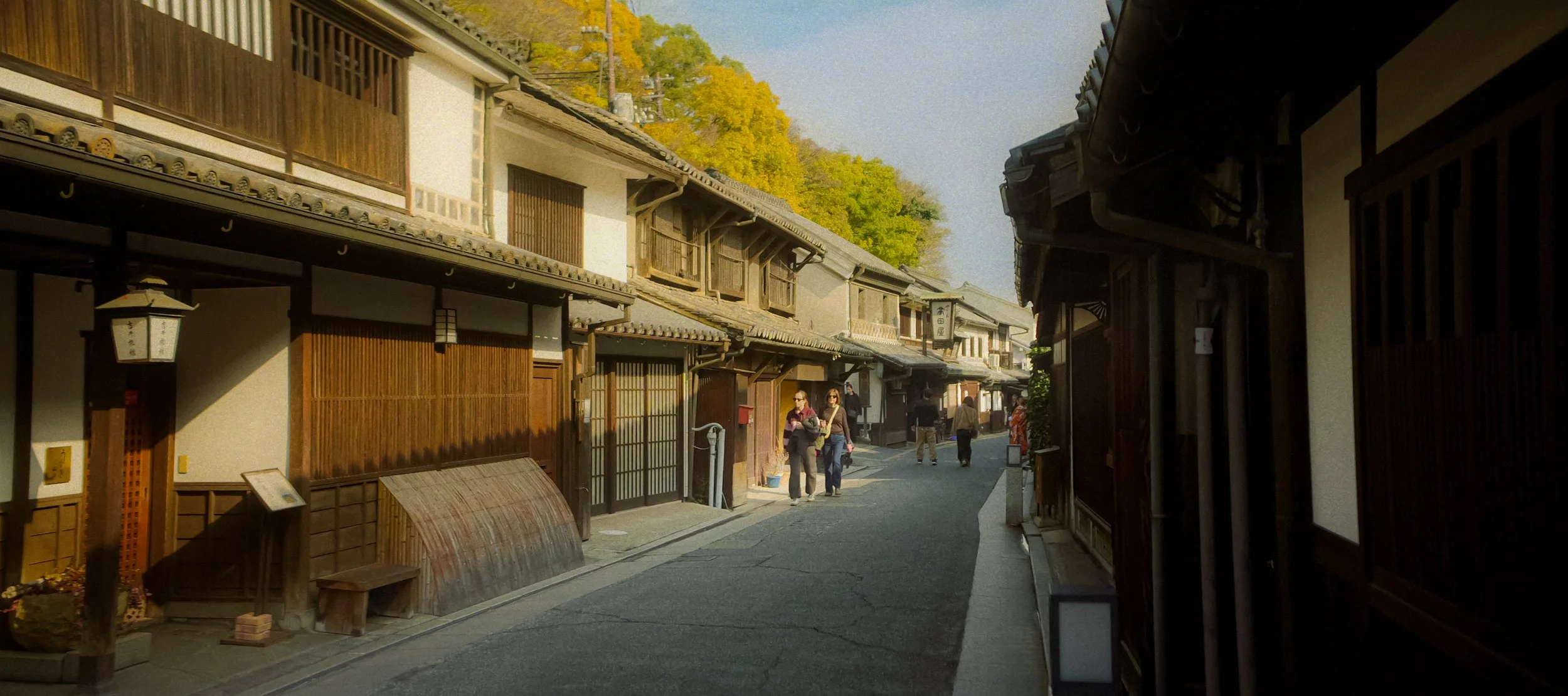 A narrow street lined with traditional Japanese wooden buildings, with a few people walking and talking on a sunny day, and trees with yellow leaves in the background.