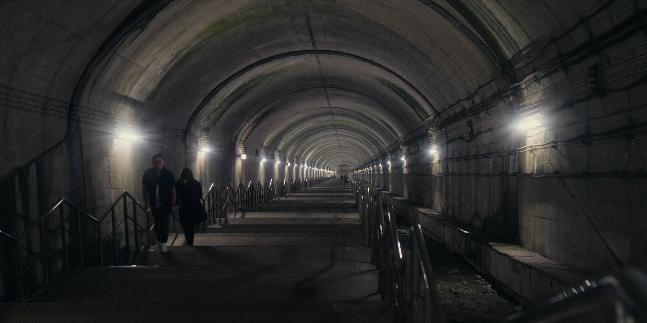 Underground tunnel with arched ceilings, illuminated by row lights along the walls, with people walking through.