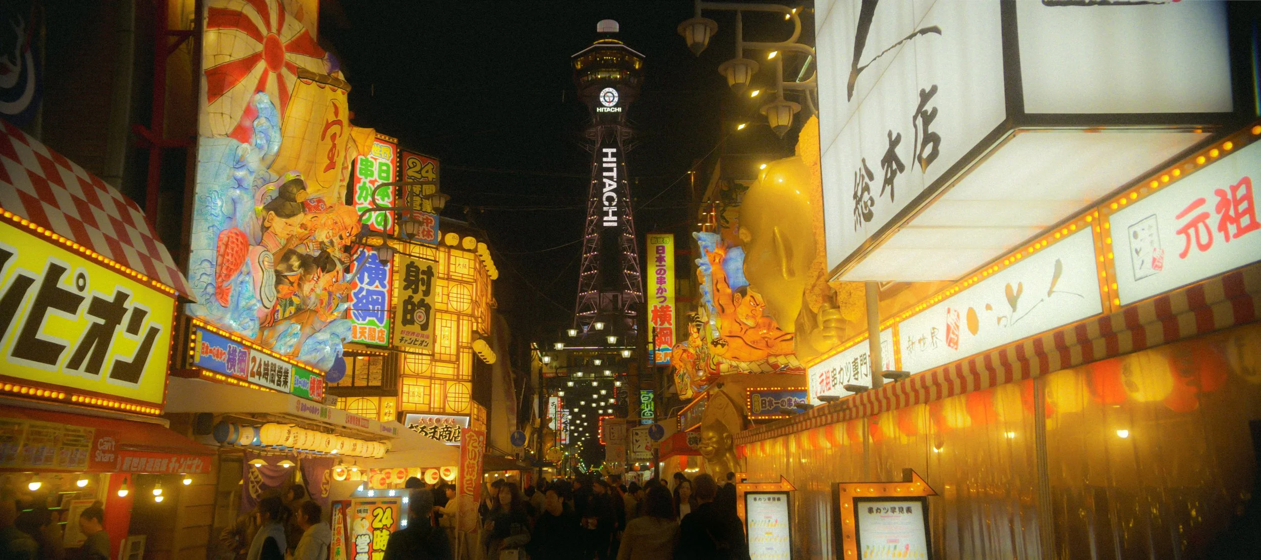 Night scene of a busy street in Japan filled with illuminated signs, colorful lanterns, and animated decorations, with a tall tower in the background.