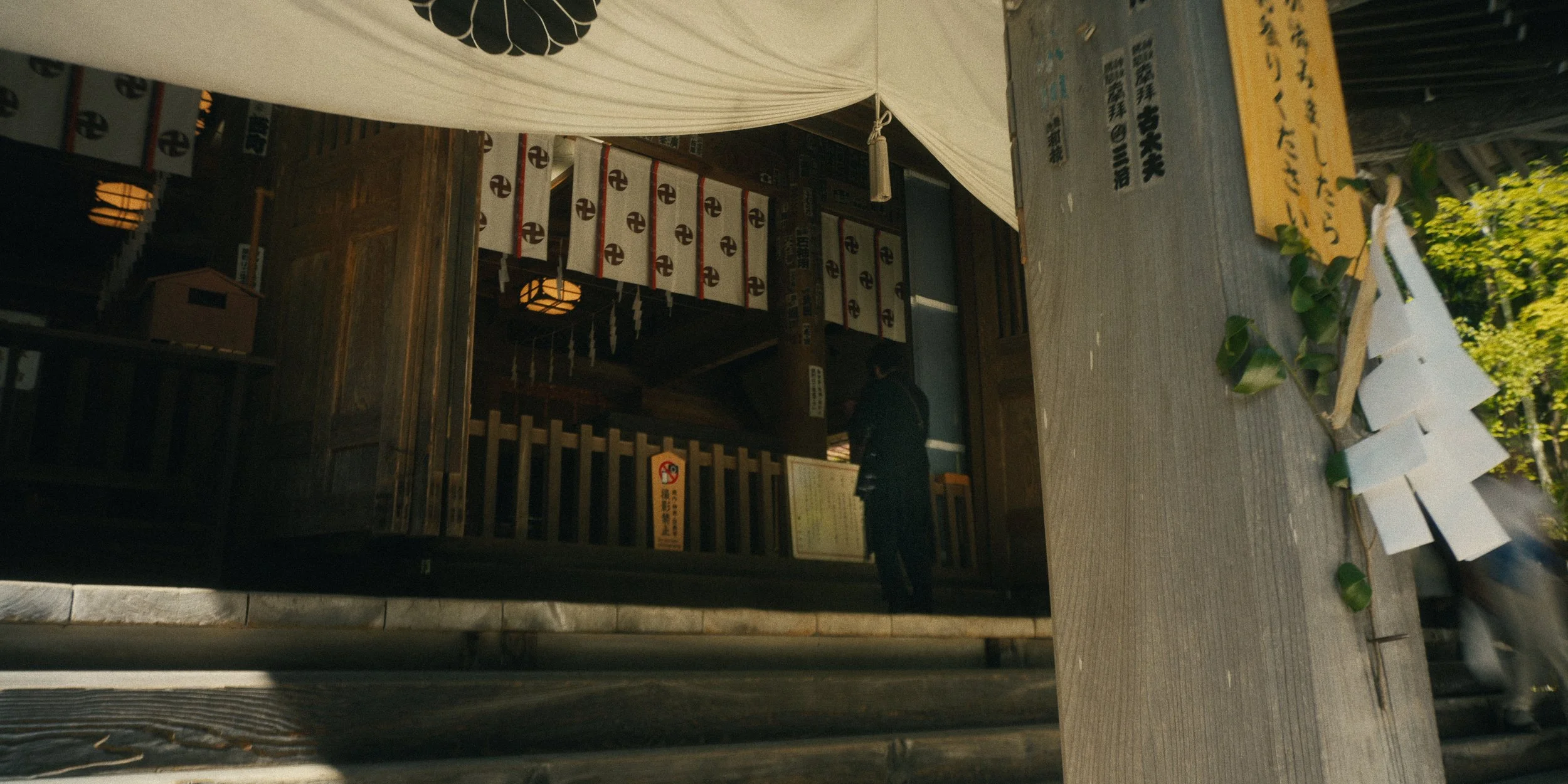 A small traditional Japanese shrine with a wooden structure, white paper lanterns, and torii gate visible. The shrine is partially covered by a white curtain and has a fence with a warning sign. There is a person standing near the entrance, and some 