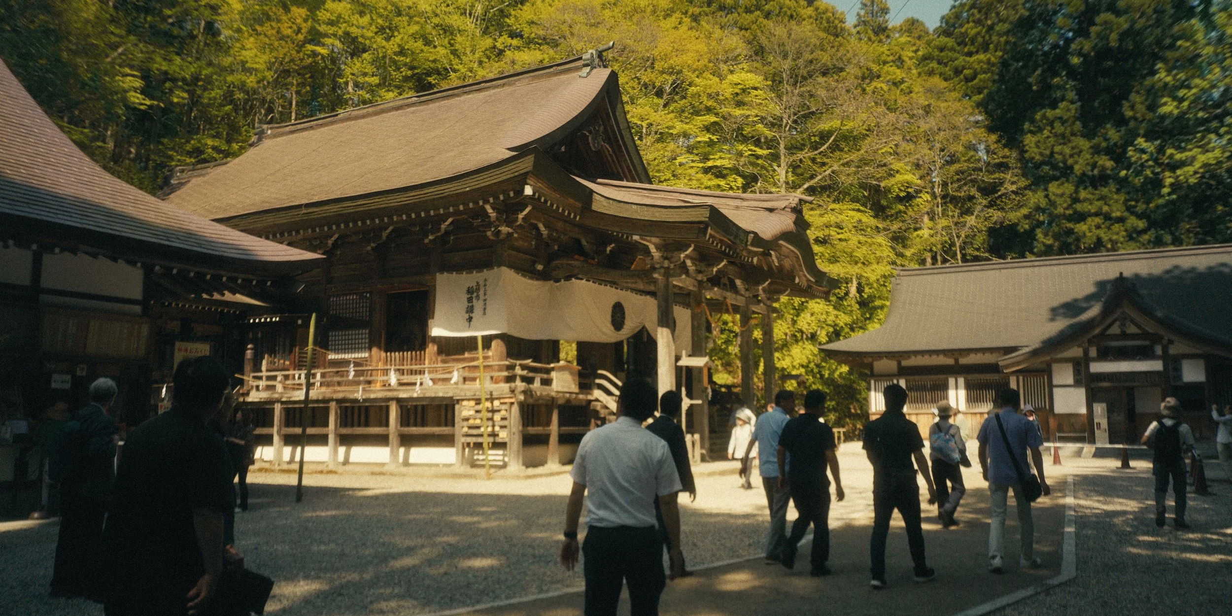 A traditional Japanese Shinto shrine surrounded by lush green trees with people walking around.