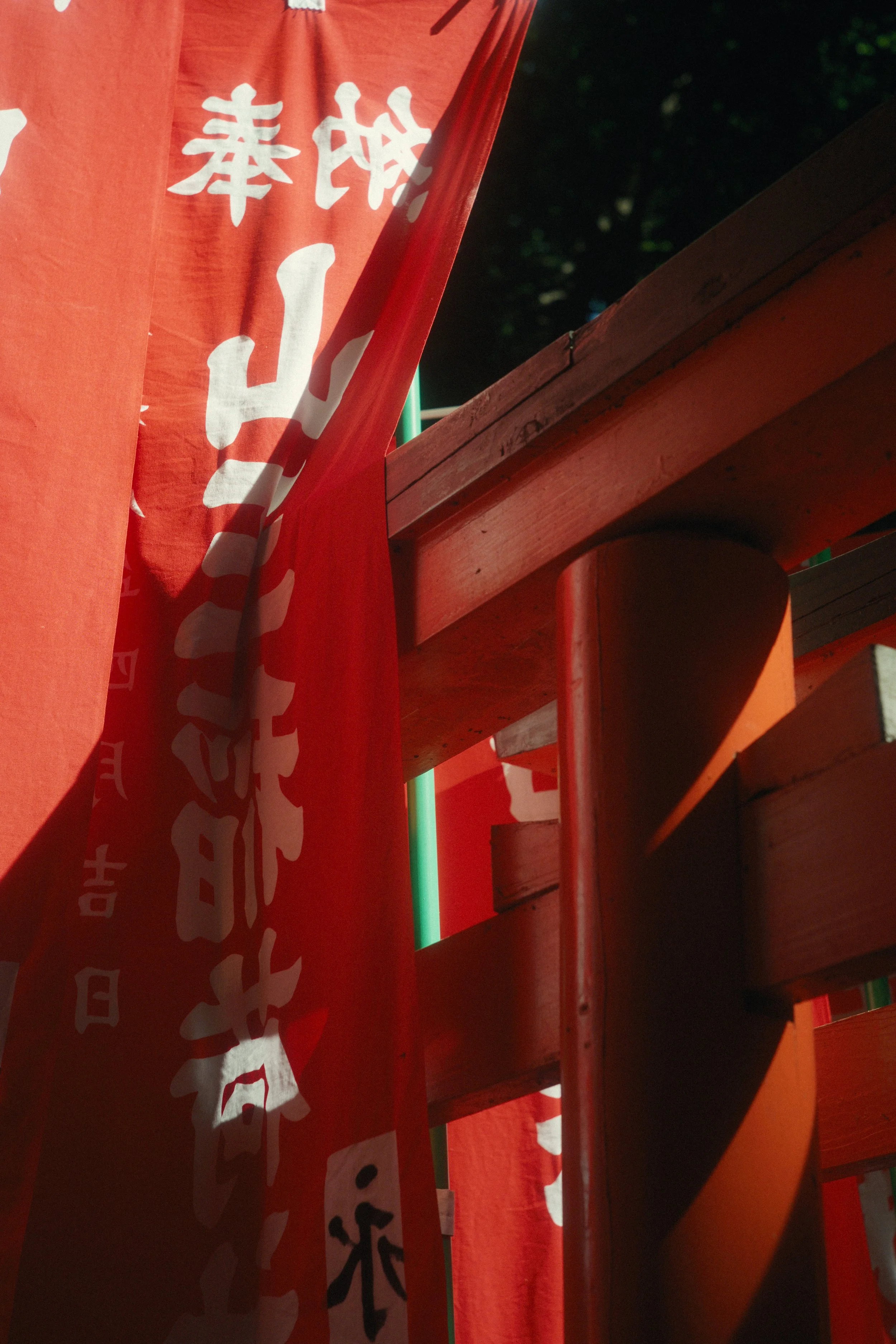 Close-up of red fabric banners with white Japanese characters, hanging next to a wooden structure, with a dark background of foliage.