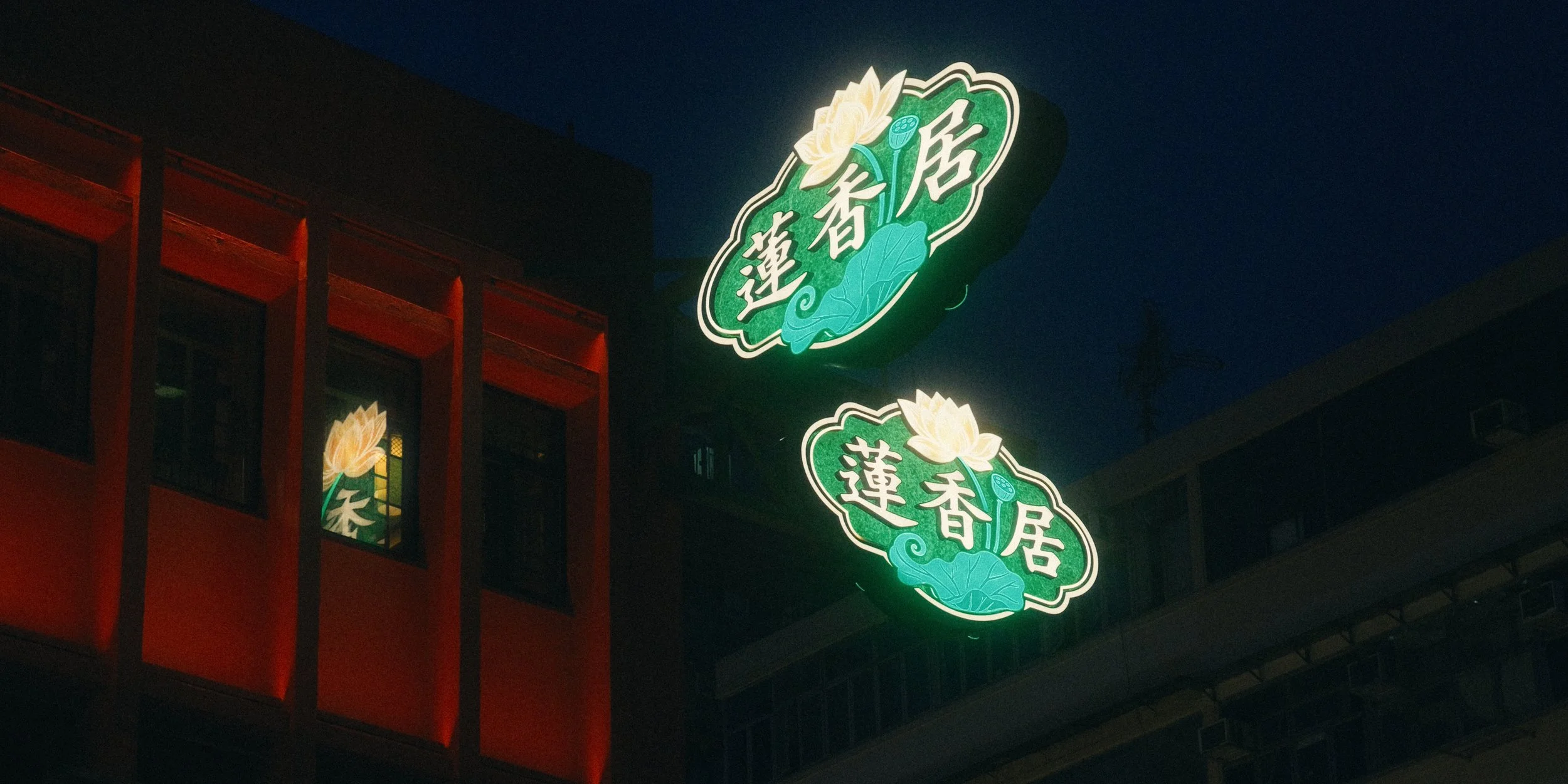 Nighttime view of illuminated green signs with Chinese characters and lotus flower graphics above a building with red accents.