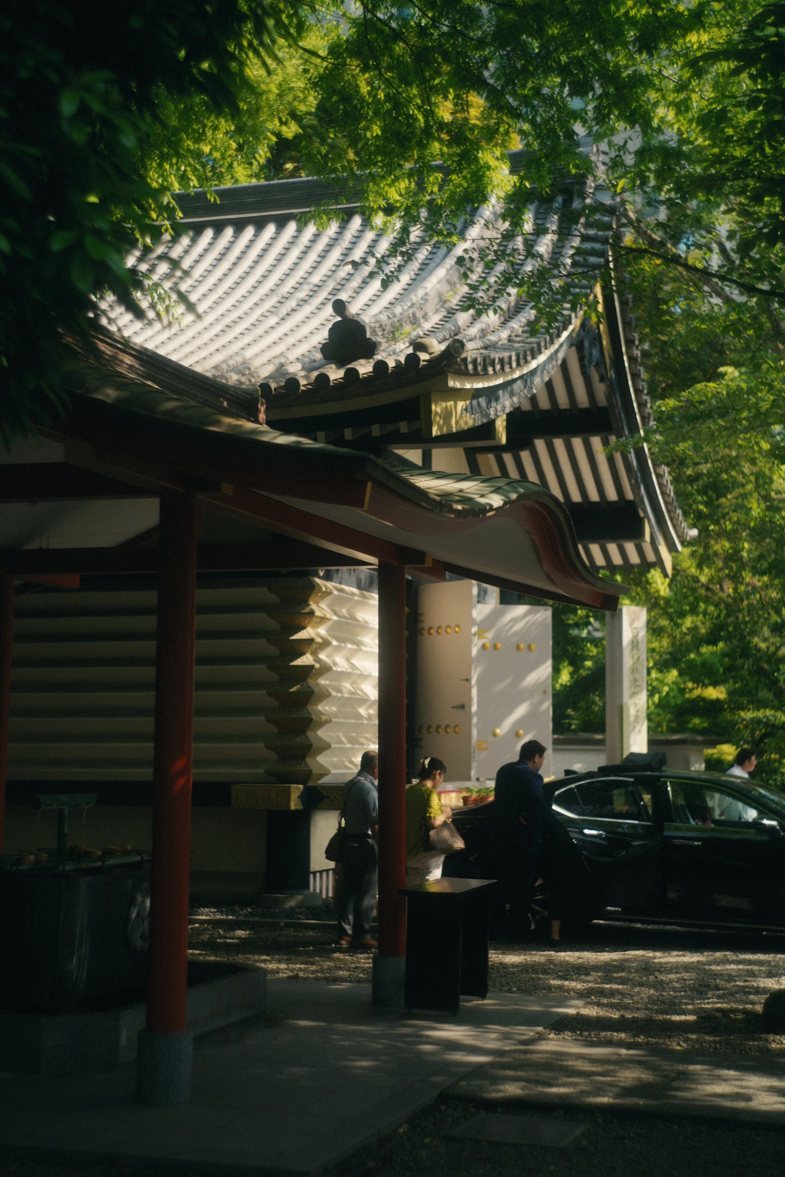 People walking and sitting outside a traditional Japanese building with a tiled roof, surrounded by greenery and trees.