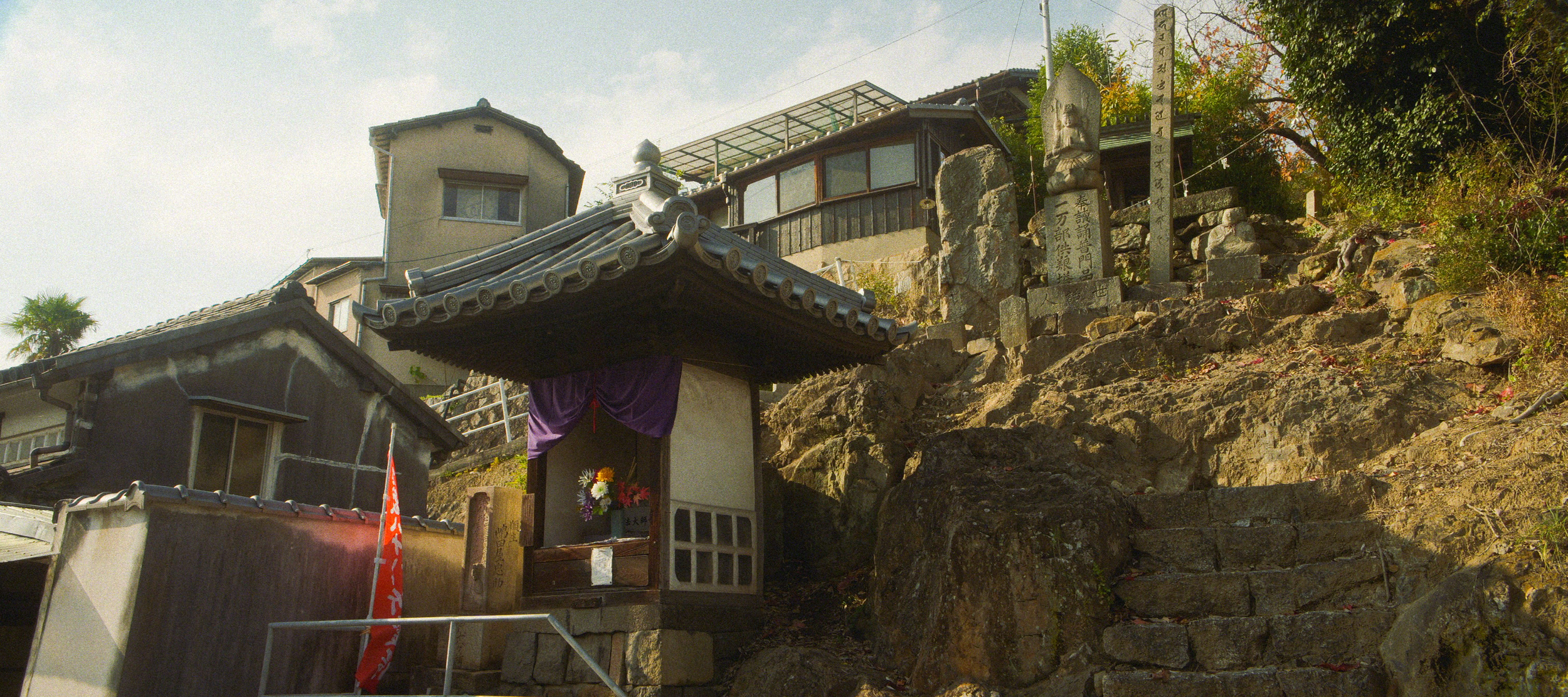 A small shrine with a tiled roof and purple curtain, situated on rocky terrain with flowers at its base. Behind it are stone statues and plaques, with houses and trees in the background.