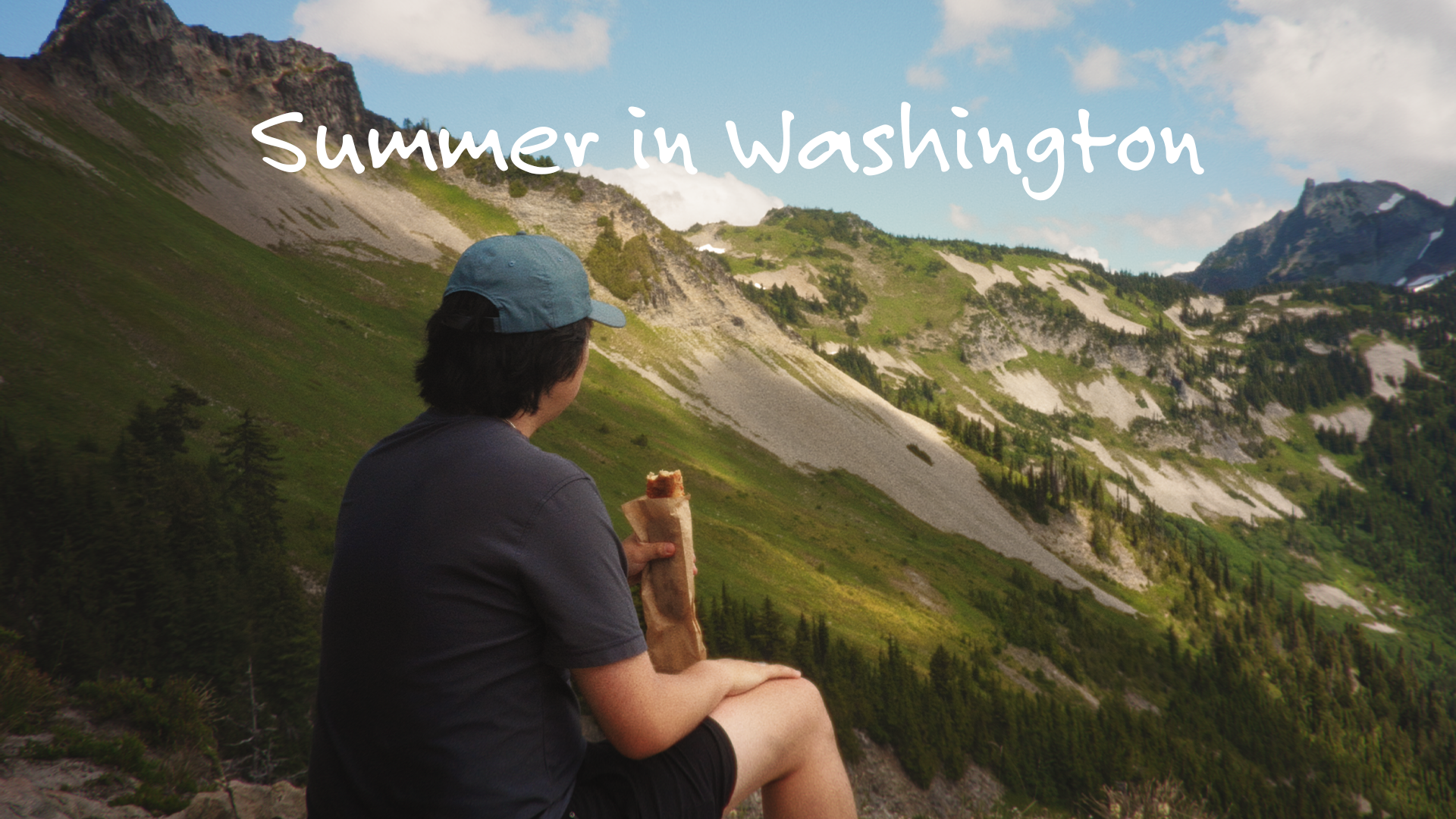 A person sitting outdoors, overlooking a mountainous landscape in Washington during summer, while eating a snack.
