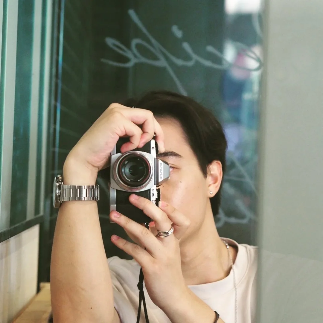 Person taking a selfie in front of a mirror with a vintage camera, wearing a white shirt, silver watch, and earrings.