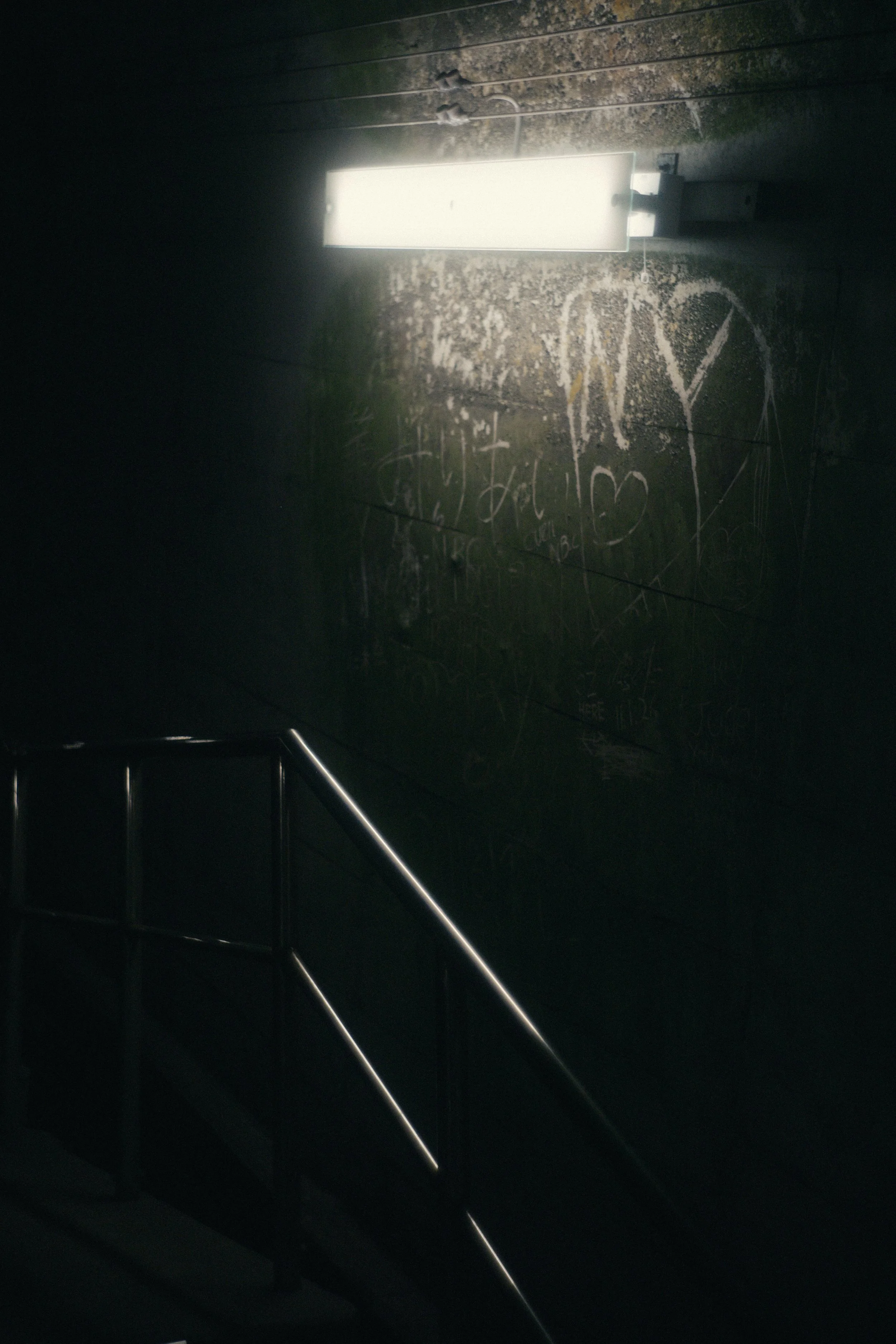 A dark staircase with metal handrails leading upwards. A wall in the background has graffiti and a runed white illuminated rectangular sign attached to a wall.
