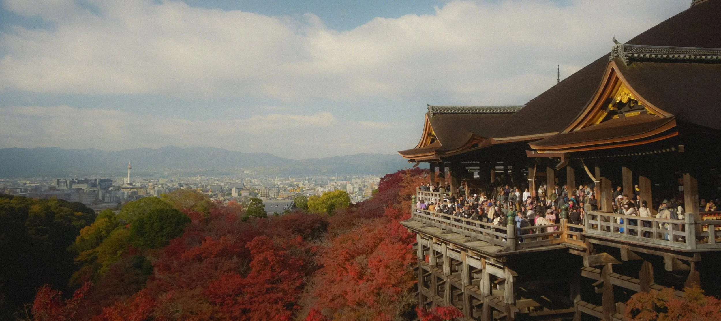 A traditional Japanese temple with a wooden structure, crowded with visitors, overlooking a cityscape with mountains in the background, surrounded by red and green autumn foliage.