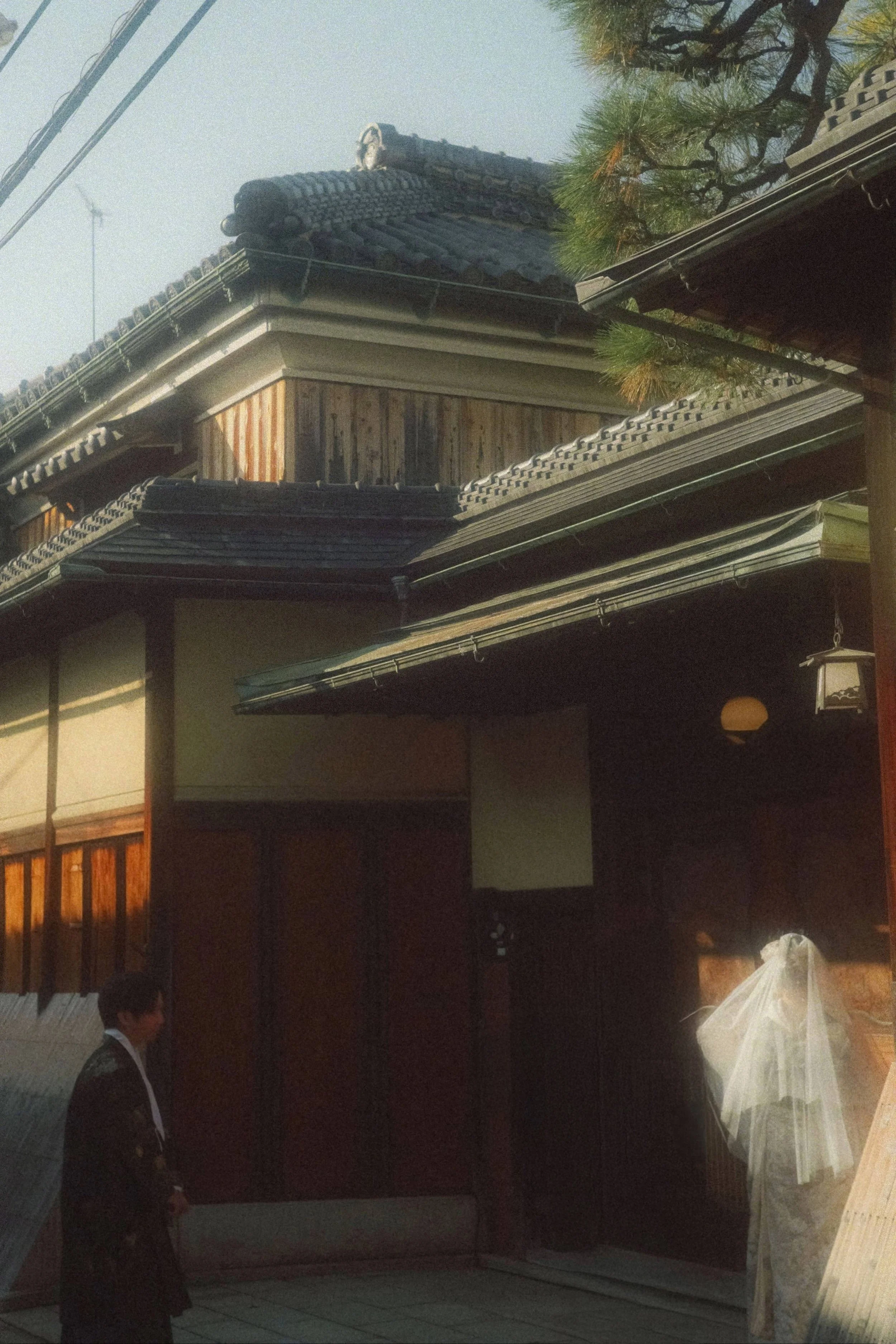Traditional Japanese building with tiled roof and greenery, with a person wearing a kimono inside.