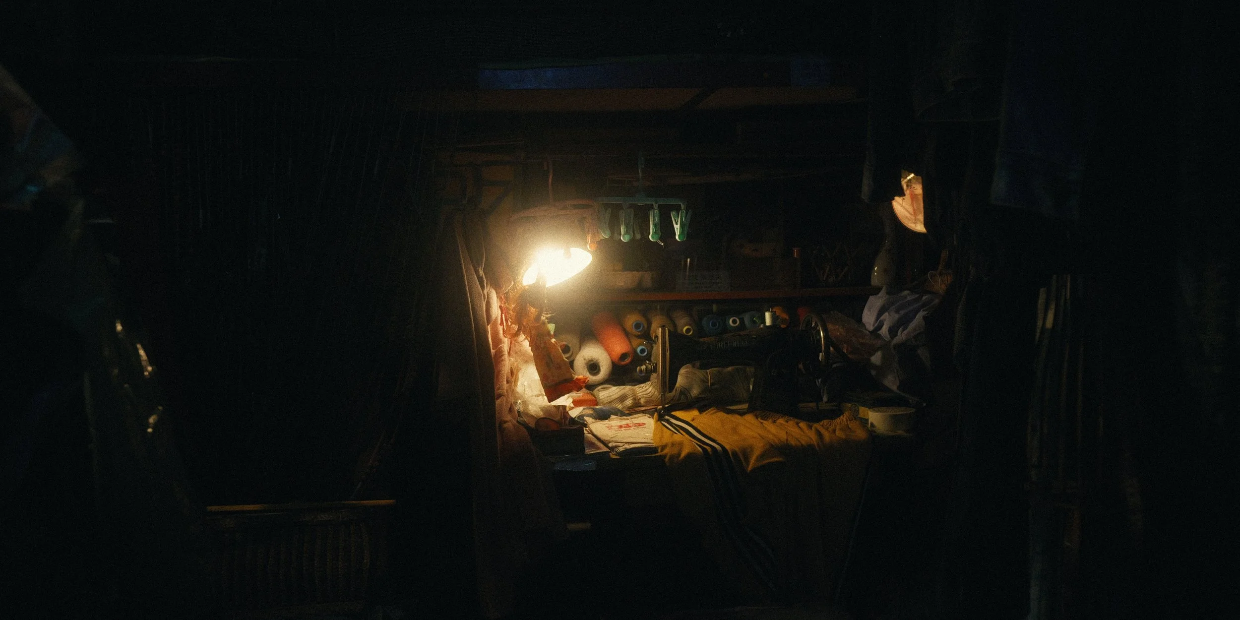 The image shows the interior of a dark room or space, illuminated by a single bright light source. Inside, there are various items such as spools of thread, possibly for sewing, hanging from the ceiling, and other craft or sewing supplies on a table 