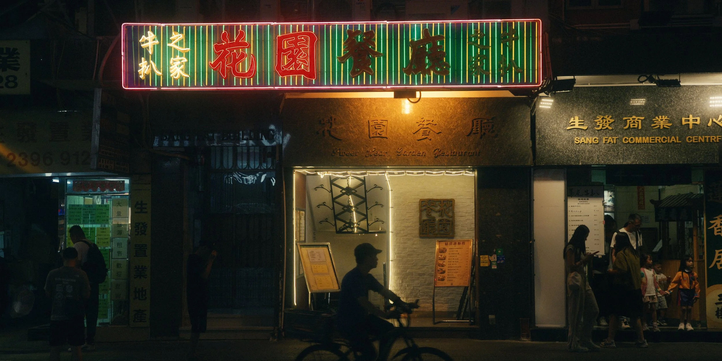 Nighttime street scene with illuminated signs in Chinese characters for a restaurant or business, with pedestrians and a cyclist visible.