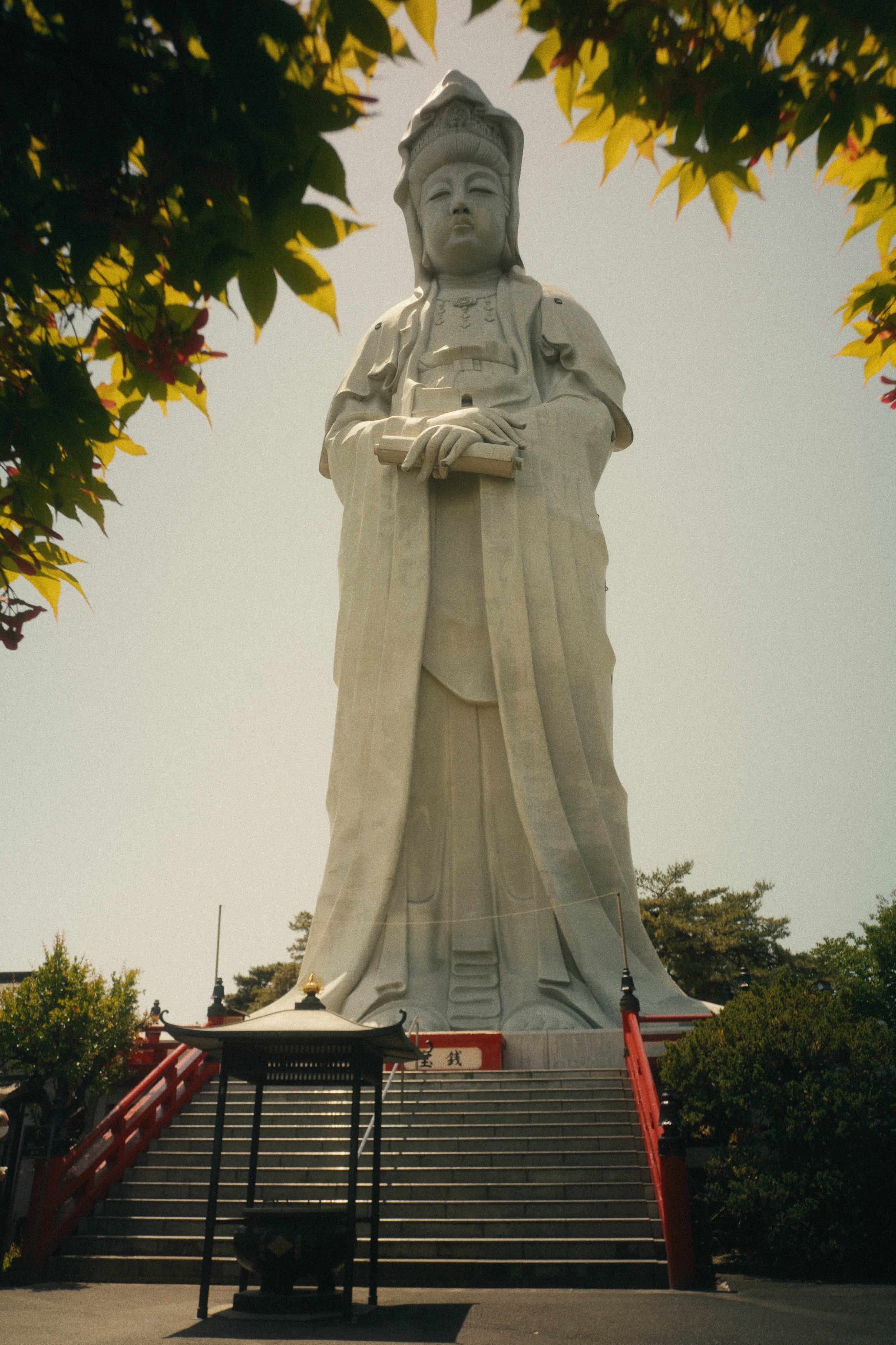 A large white statue of a historical warrior figure stands at the top of a staircase with red railings, surrounded by green trees under a cloudy sky.
