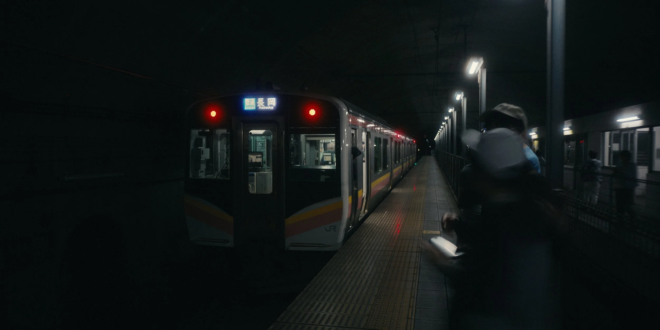 An underground train station with a train stopped at the platform. The station is dimly lit with overhead lights, and a few passengers are visible on the platform.